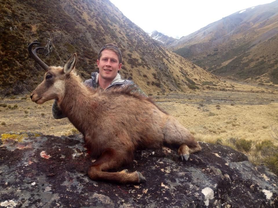 Man posing with a dead chamois he's holding on a rock. They are in a mountain setting, with brown and green slopes in the background.