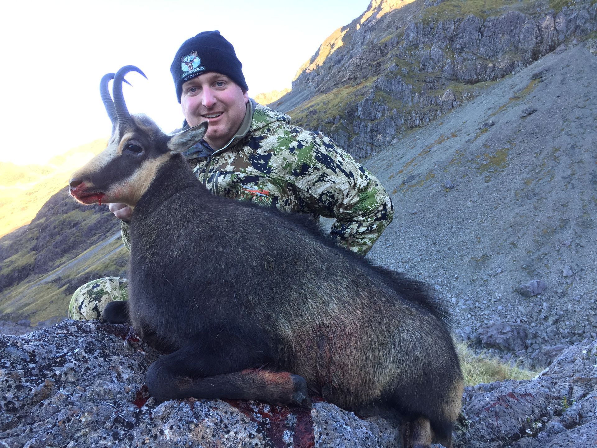 Hunter in camouflage with a dead chamois. They're posing on rocks in a mountainous landscape, the chamois's dark fur and horns are prominent.