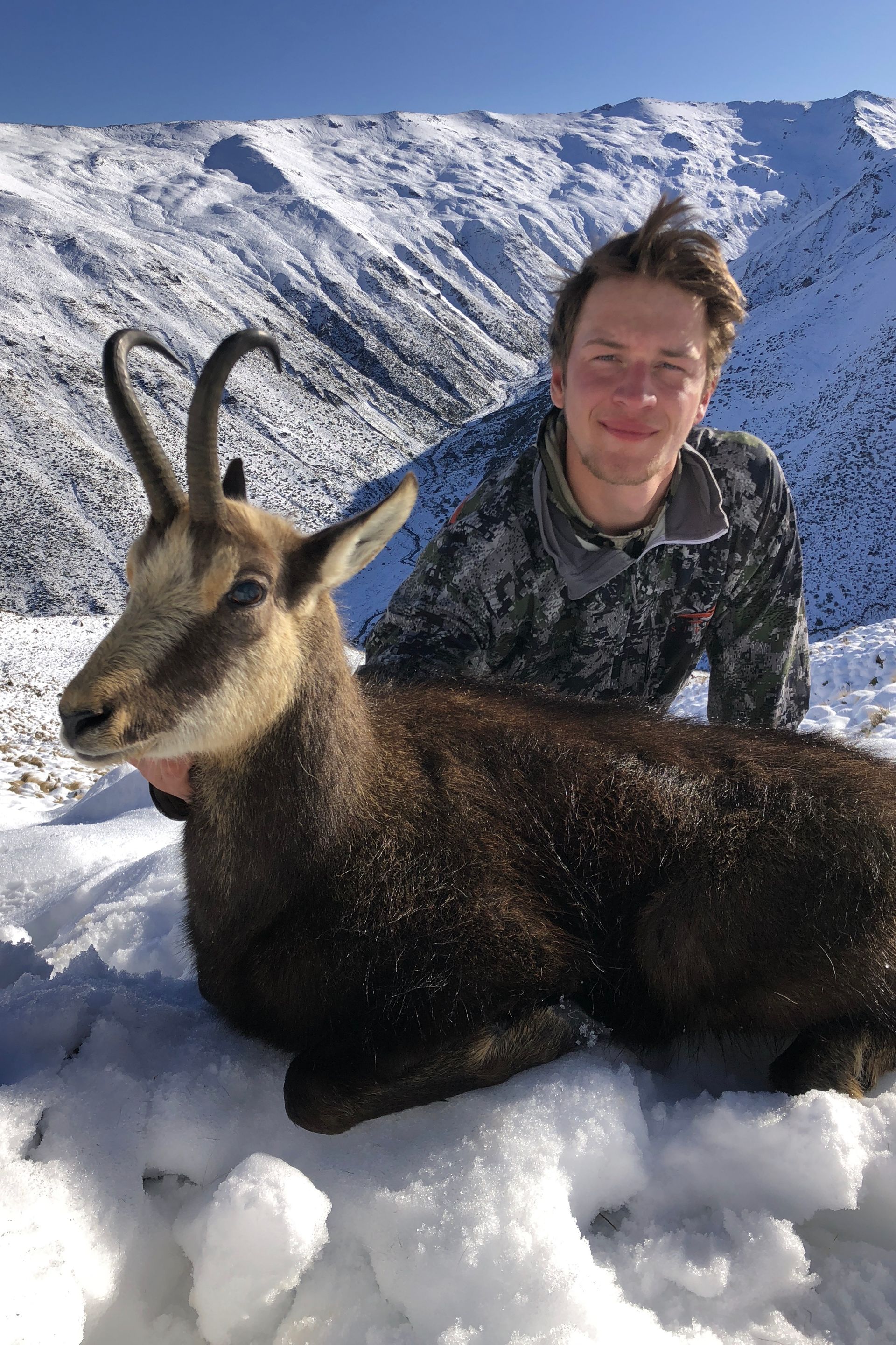 A young man kneels next to a harvested chamois in a snowy mountain setting. Both are covered in snow; the man smiles.