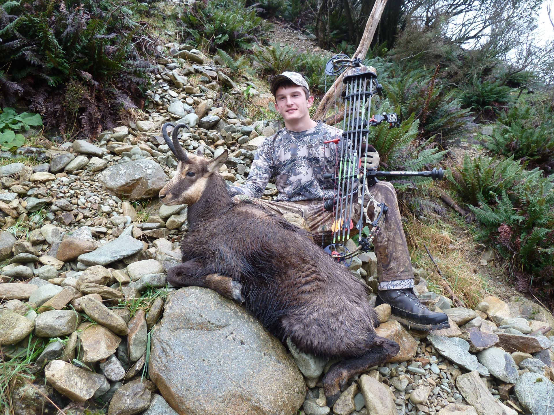 A young hunter in camouflage sits next to a recently hunted chamois and bow on a rocky hillside.