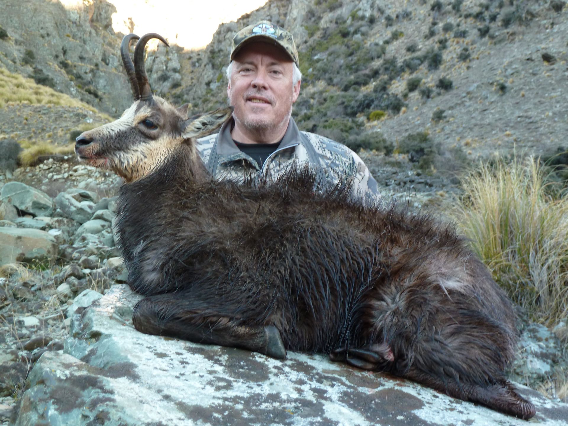 A man in camouflage hat and jacket poses with a large, dark-furred chamois he has hunted in a rocky, mountainous area.