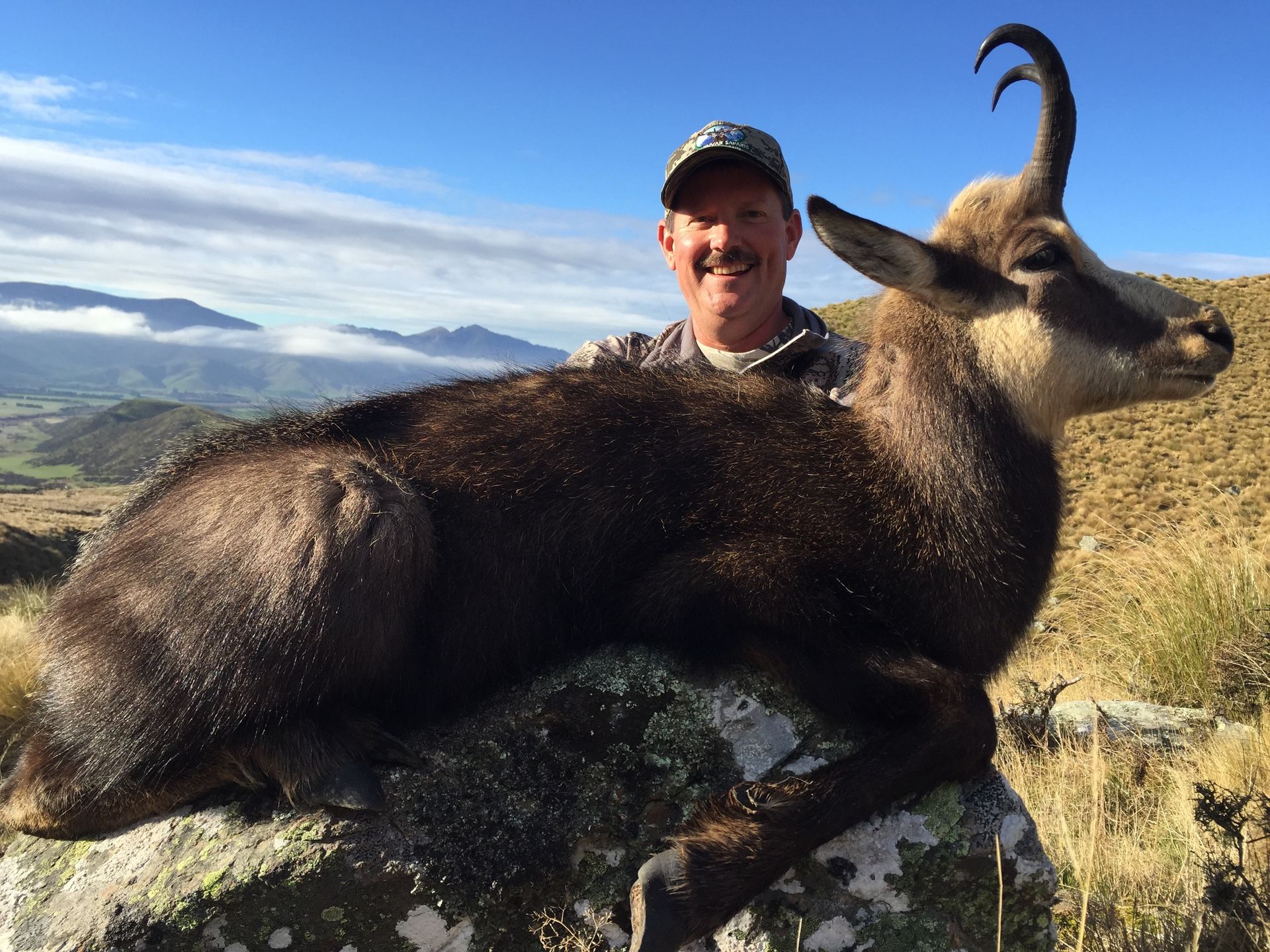 Man smiles, posing with a dead chamois on a rock in a mountain landscape. The animal has curved horns and dark fur.