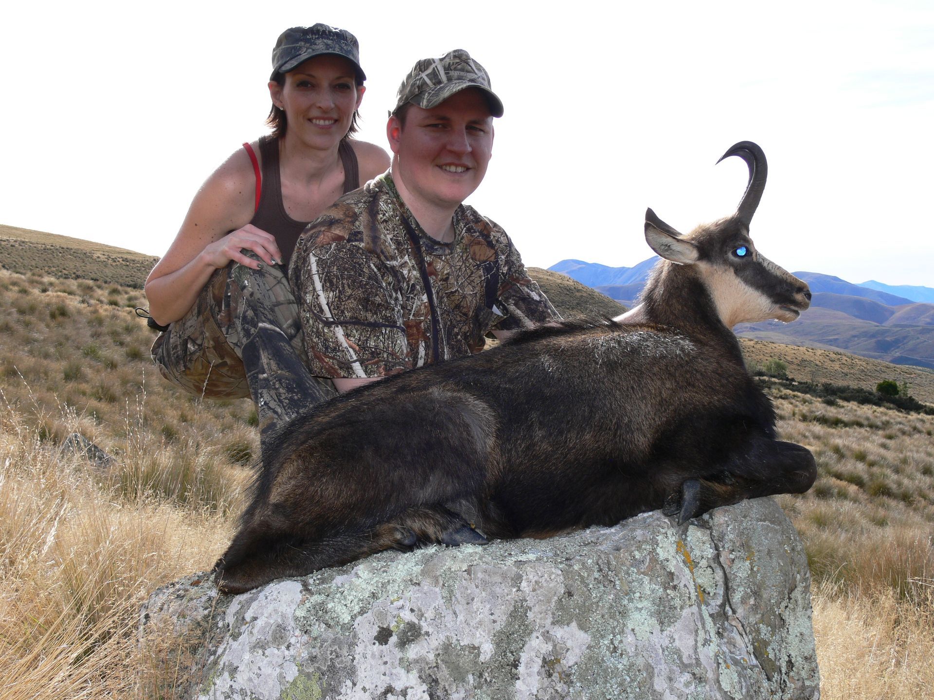 A man and woman in camouflage pose with a dead chamois on a rock, in a dry, hilly landscape.
