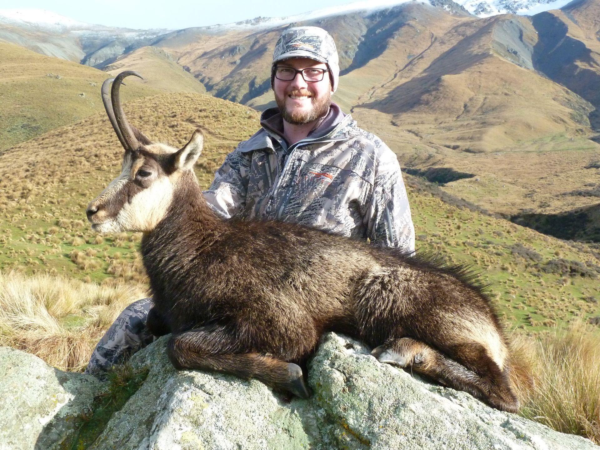 Man in camouflage holding a dead chamois with curved horns on a rocky outcrop, mountains in the background.