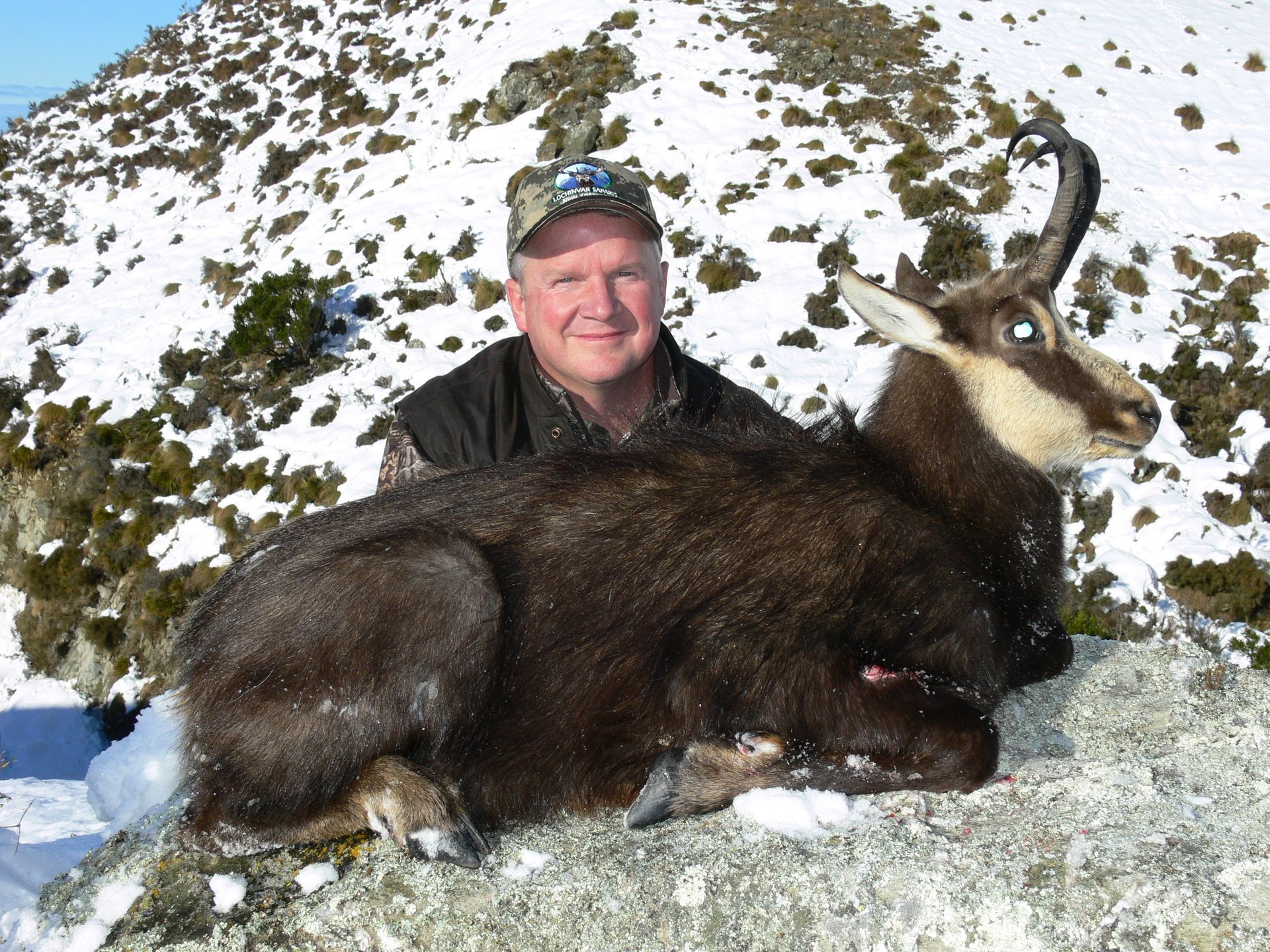 Man in camo hat smiles, holding a chamois he hunted, on a snowy mountain.