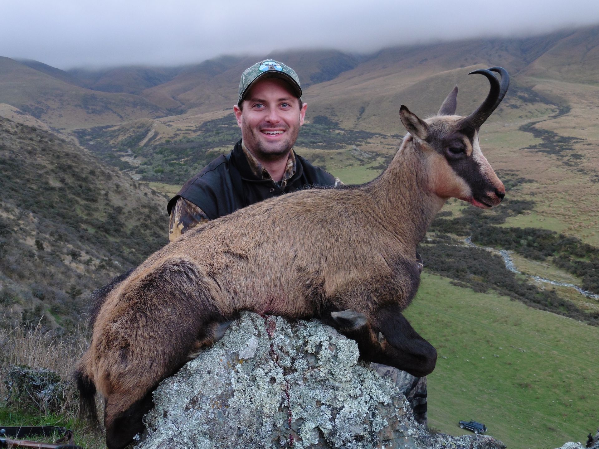 Man smiles, holding a chamois he's hunted in a mountainous, grassy landscape. The animal is tan and black with curved horns.