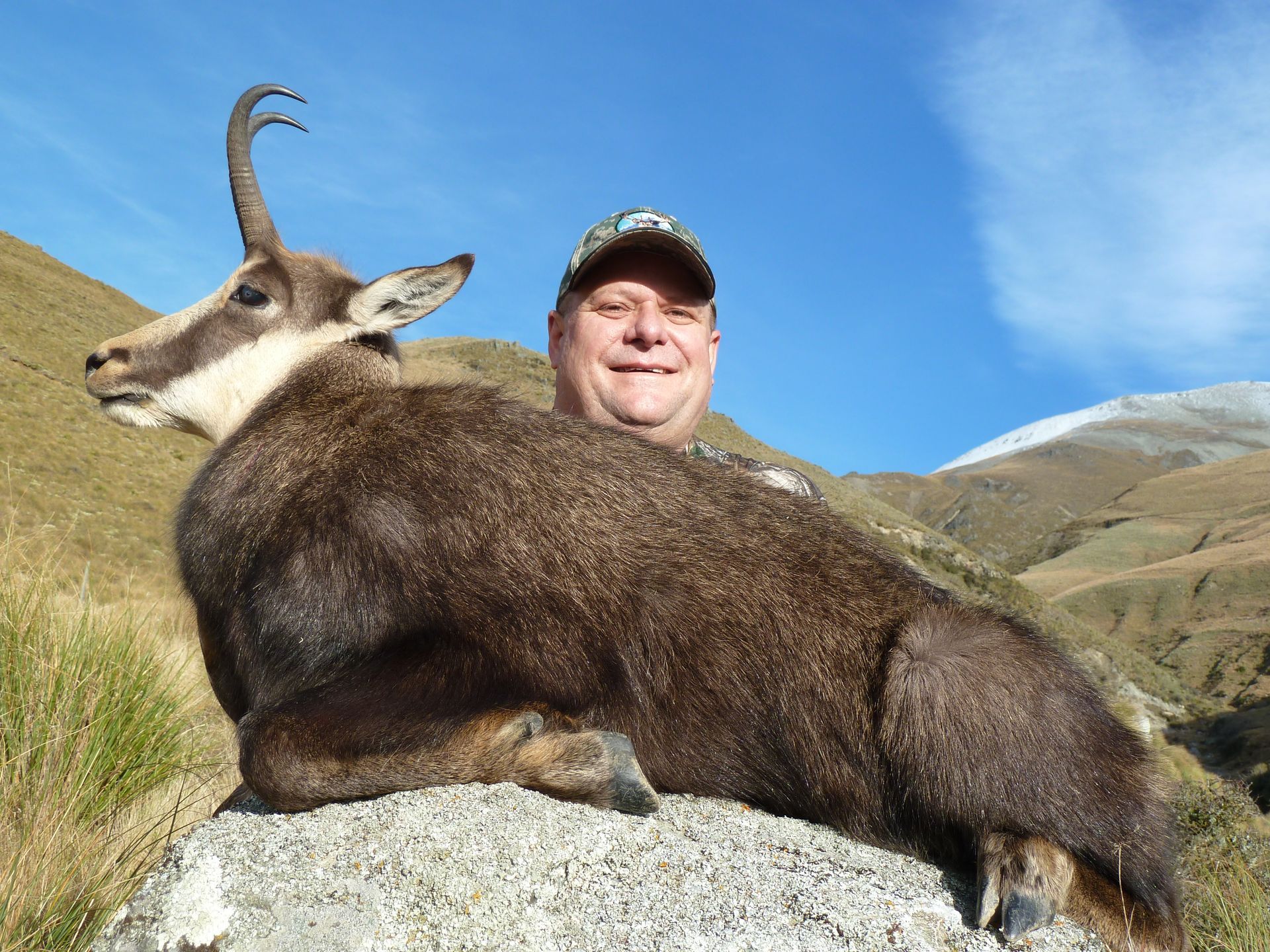 Man posing with a dead chamois goat on a rock in a mountain landscape. The man is smiling. Blue sky in the background.