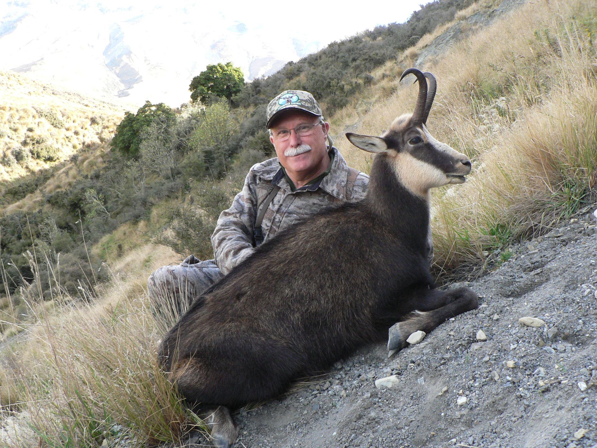 Hunter kneeling beside a freshly-hunted chamois on a hillside. The chamois is dark-furred with curved horns, and the hunter wears camouflage.
