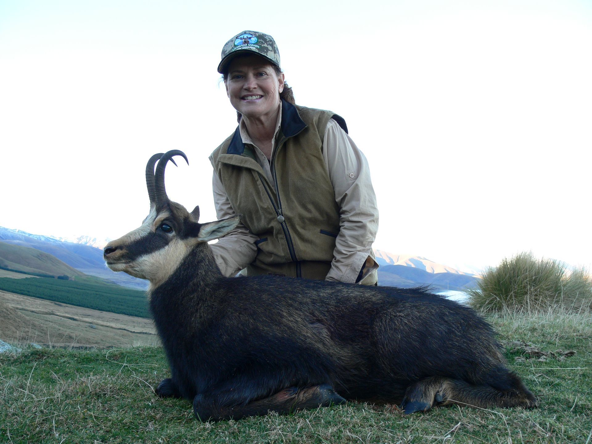 Woman in hunting attire smiles beside a harvested chamois in a grassy field, mountains in the background.