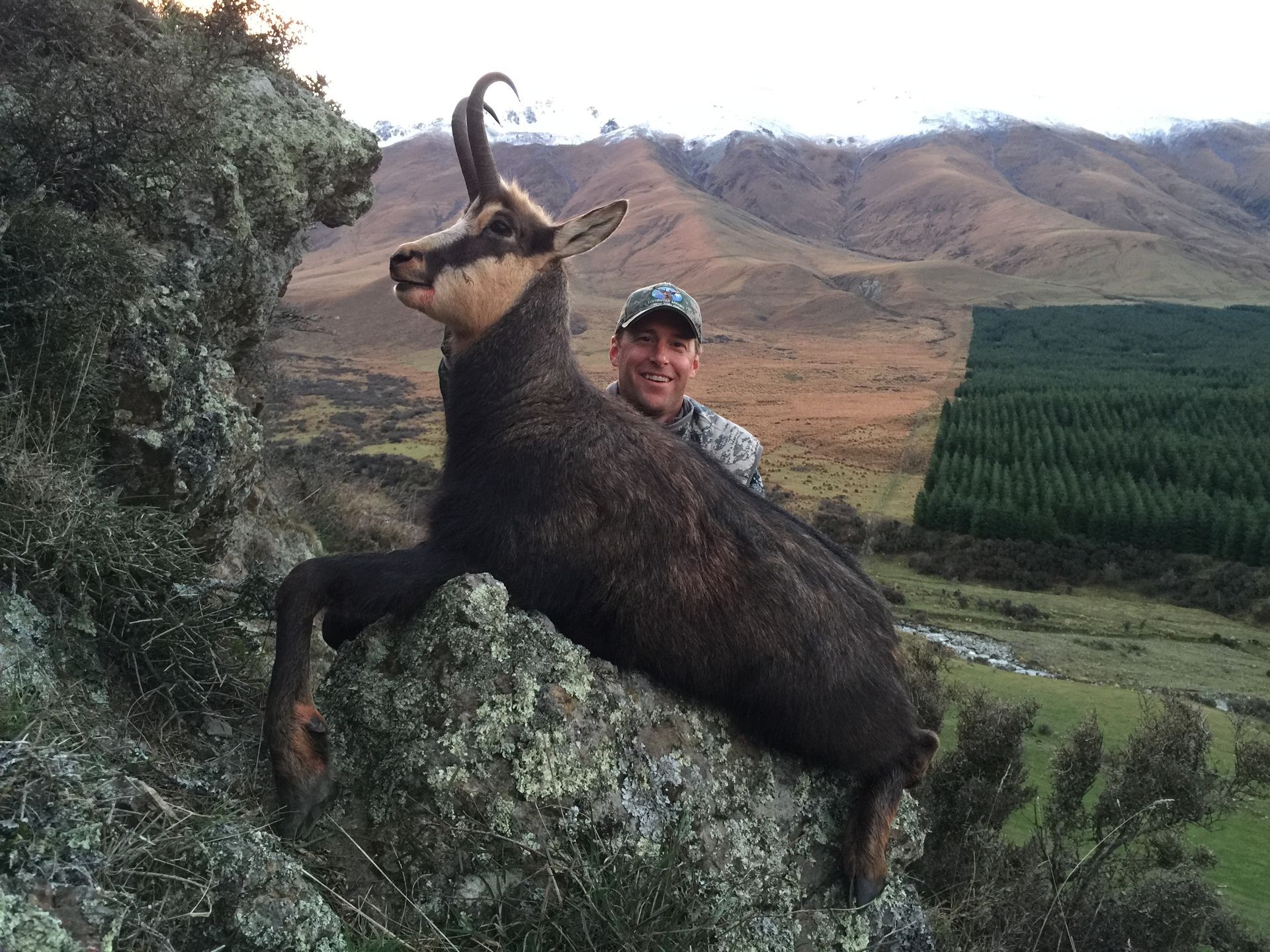 Man smiles, holding a chamois he hunted on a rocky ledge with a mountain backdrop.