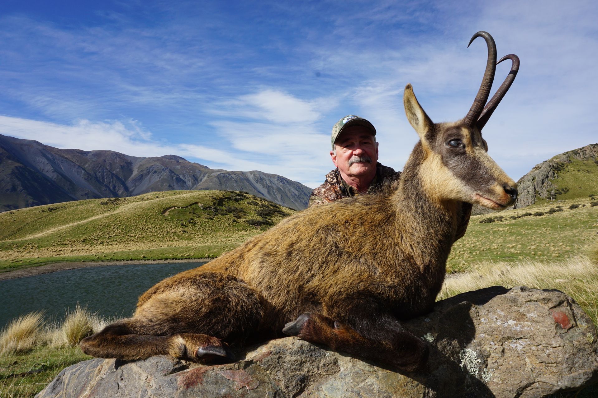 Man in camouflage with a dead chamois, posed on a rock in a mountain landscape. The chamois has large curved horns.