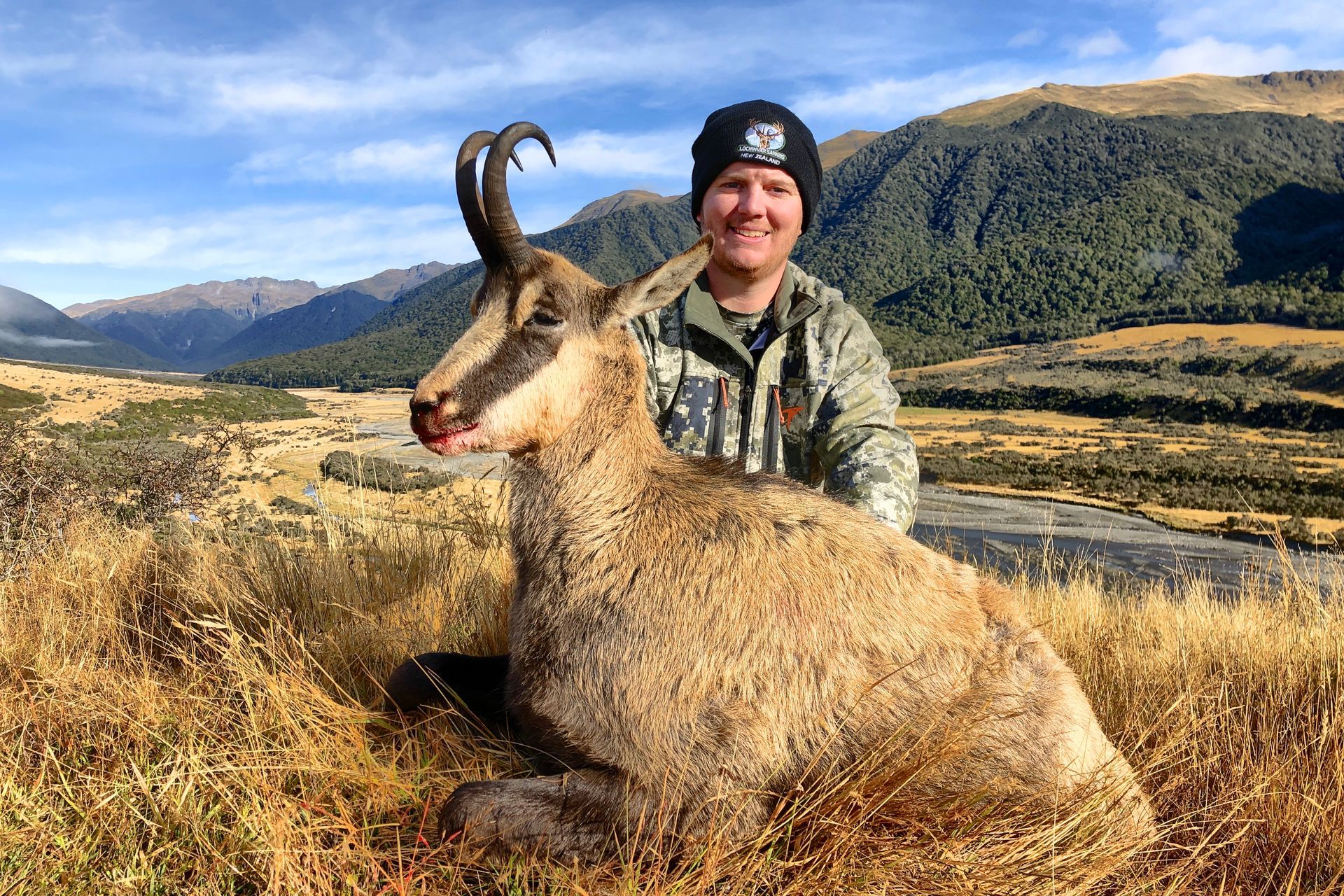 Man in camouflage with a dead chamois in a mountainous field. The chamois has large horns; the man is smiling.
