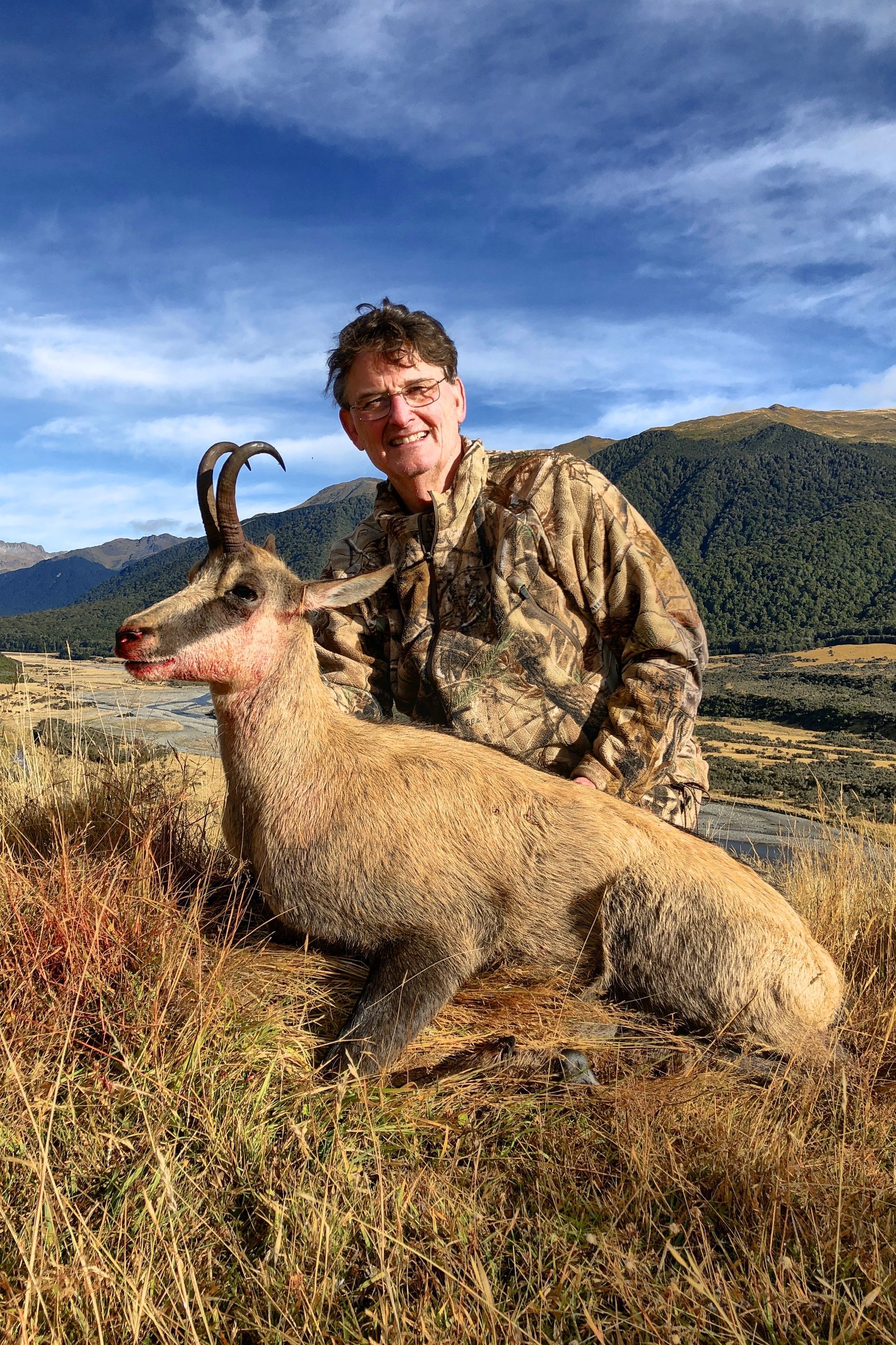 Man in camouflage with a chamois he has hunted. Blue sky and mountains in the background.