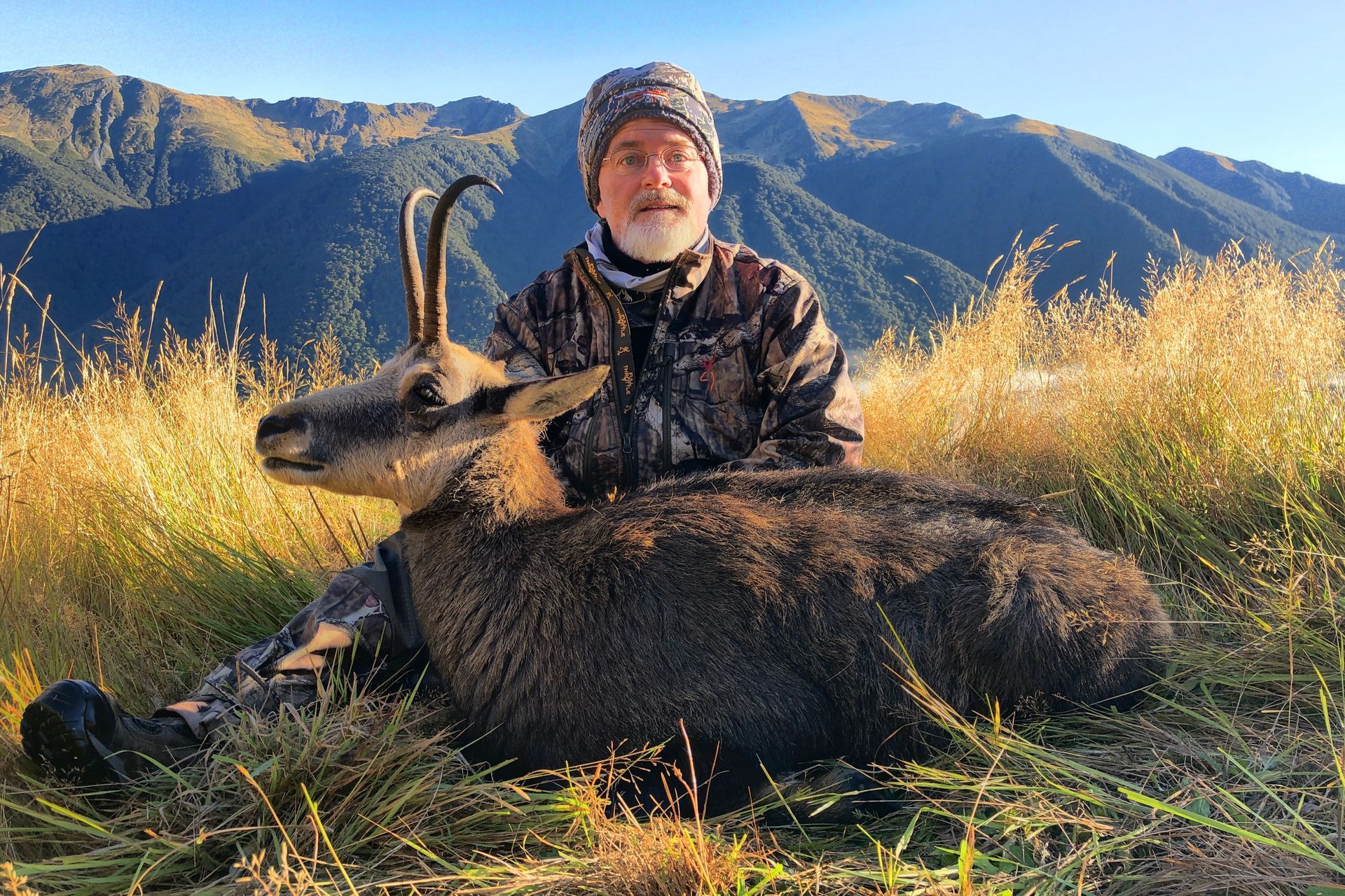 A man in camouflage, smiling, sits next to a dark-furred chamois in a grassy field, mountains in the background.