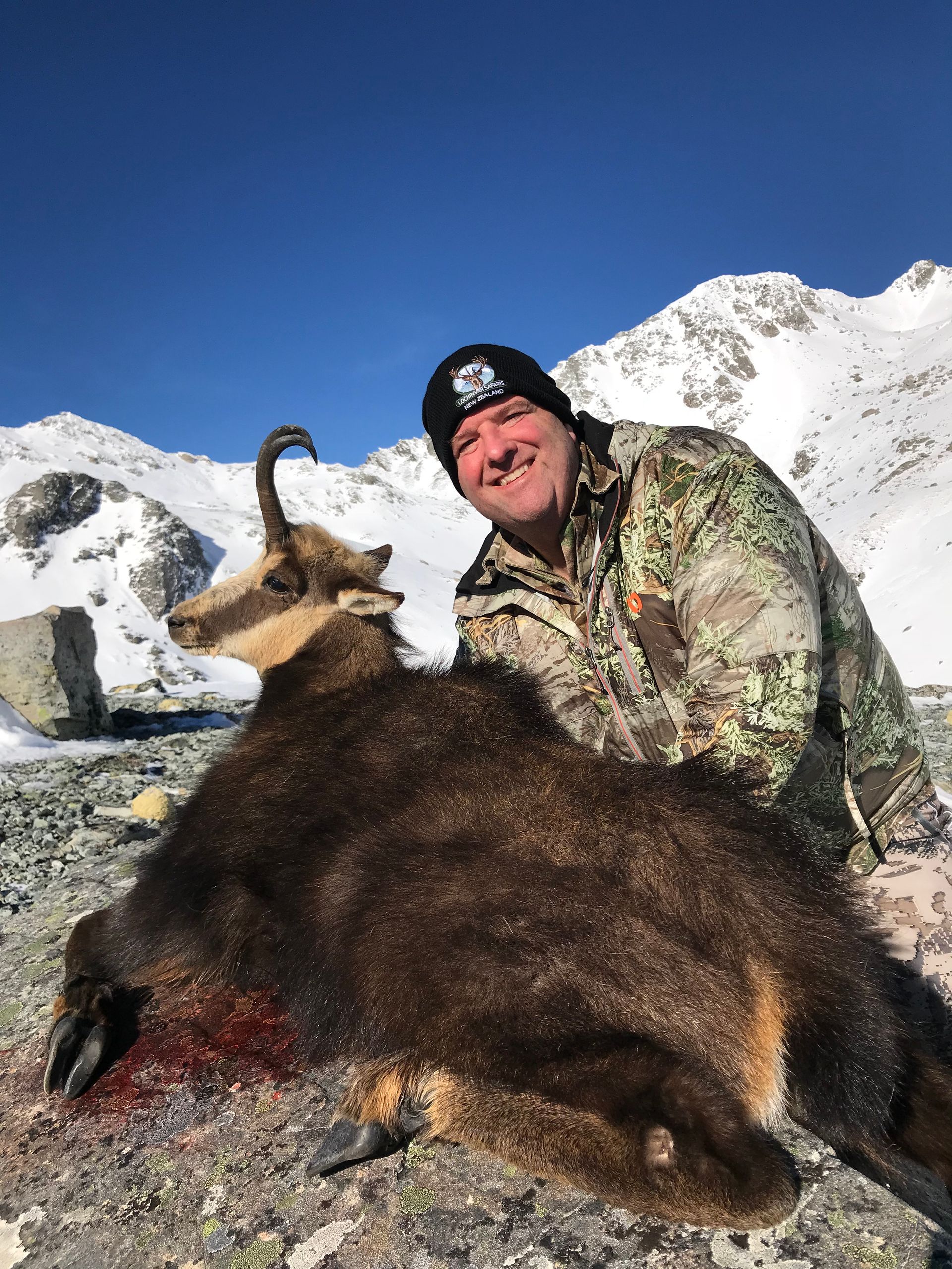 Man in camouflage jacket smiles next to a dead chamois in a snowy mountain setting under a clear blue sky.