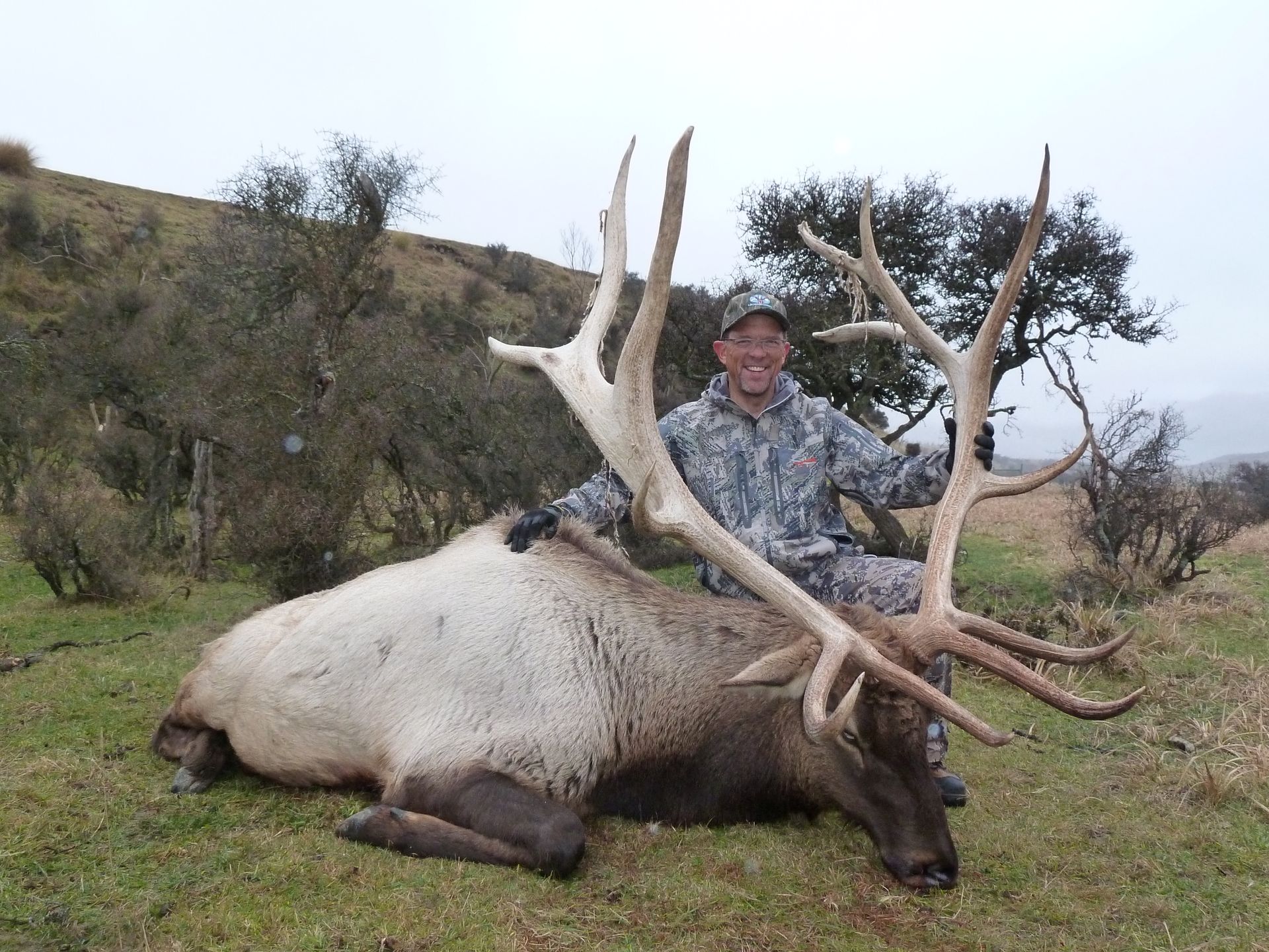 Man in camouflage smiles next to a large elk with impressive antlers on a grassy hillside.