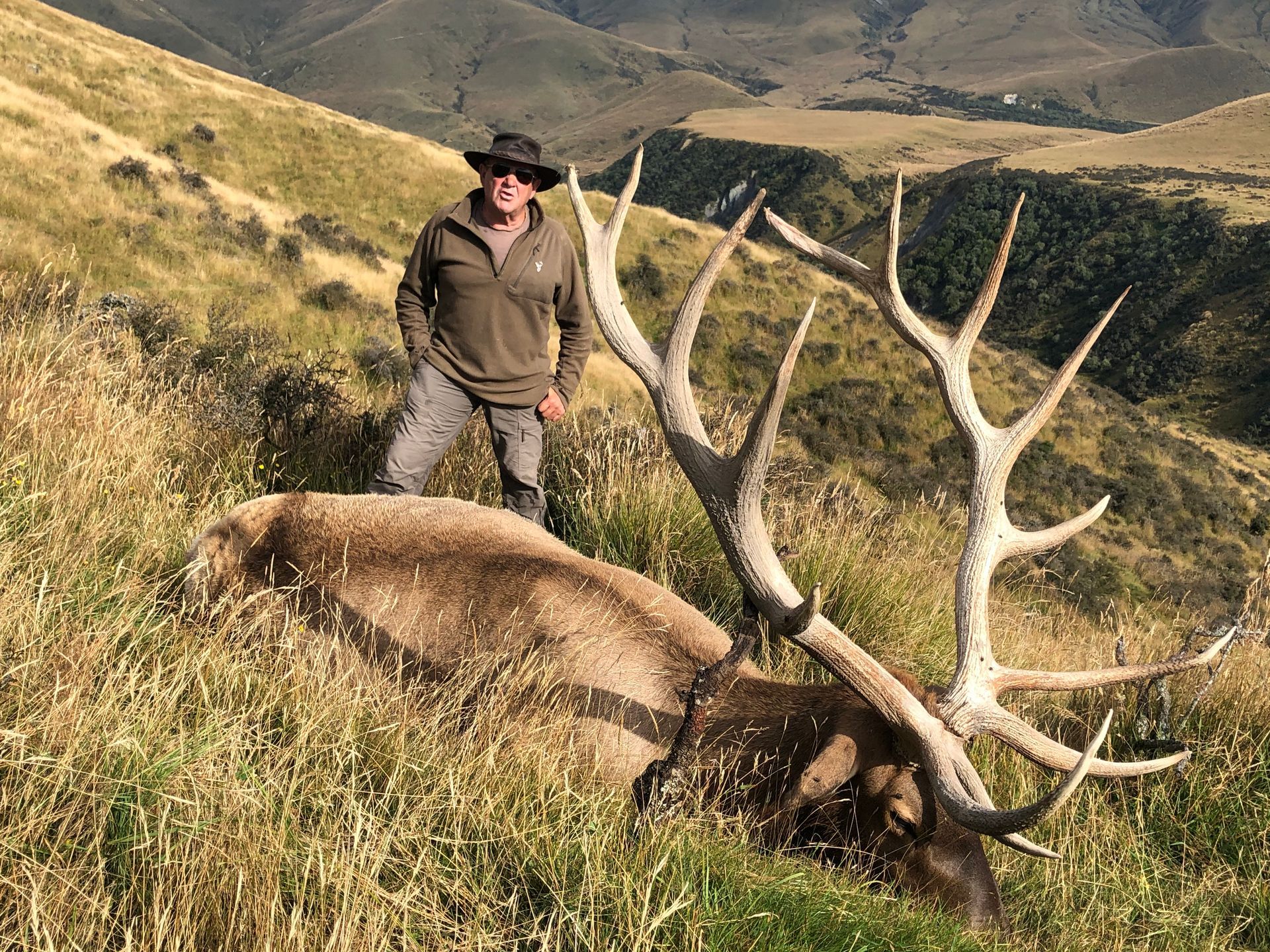 Man in hunting gear stands beside a large dead elk in grassy hills. He is smiling, wearing a hat, and the elk has impressive antlers.