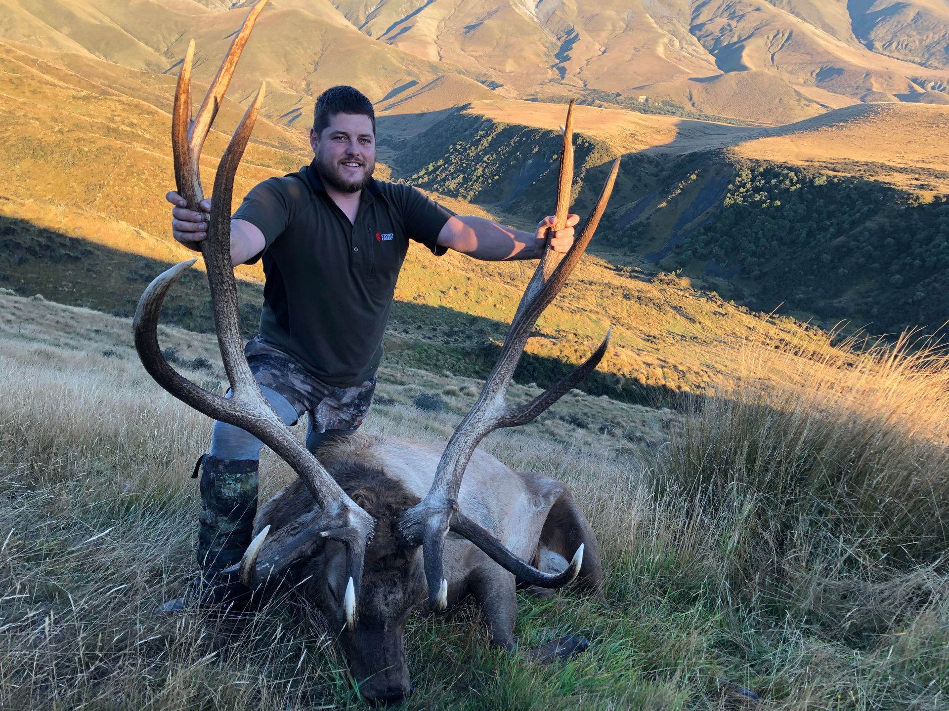 Man posing with a large elk he has hunted in a mountainous, grassy landscape. The man wears hunting attire and smiles.