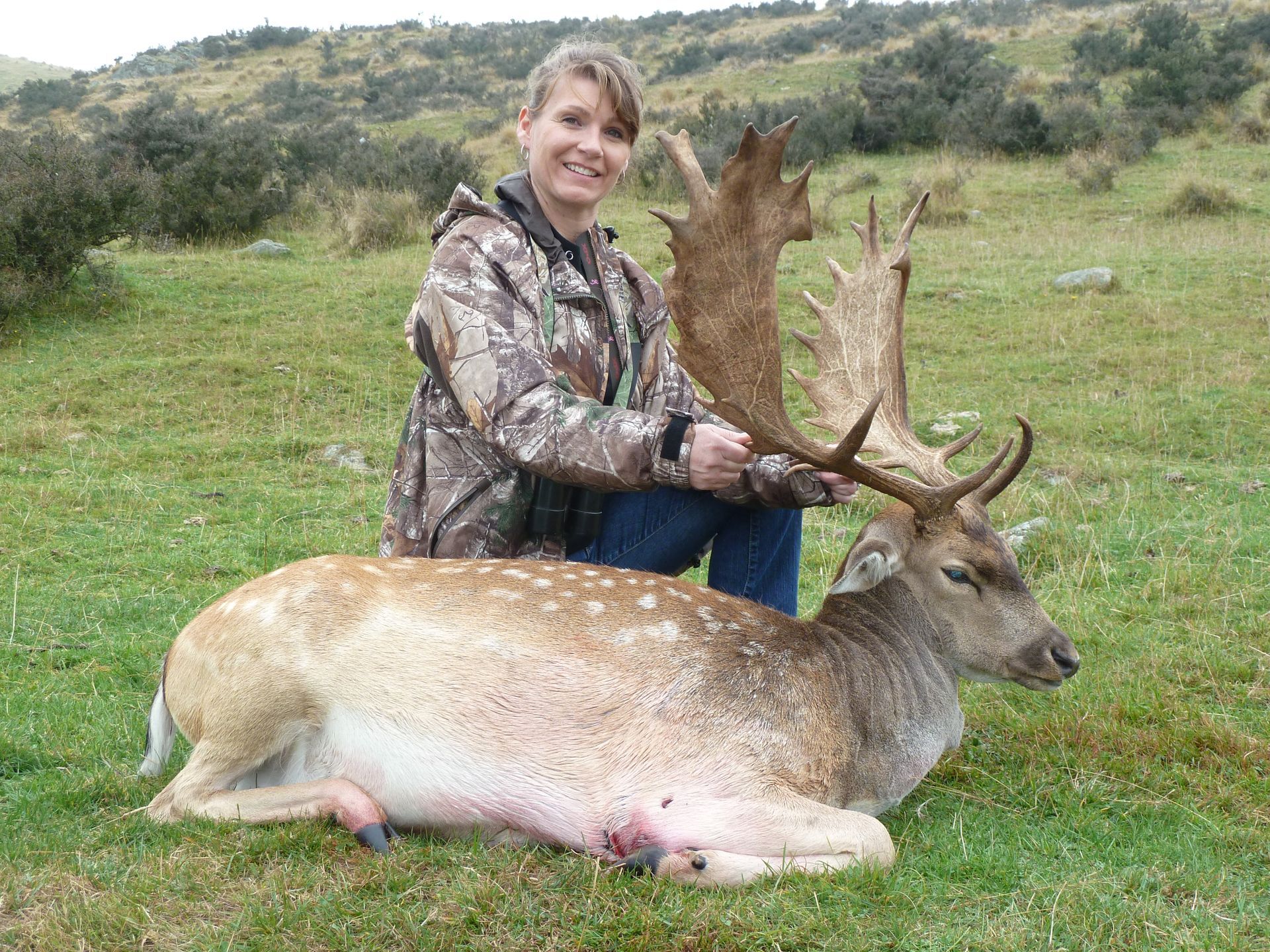 Woman in camouflage kneeling beside a felled deer with large antlers in a grassy field, smiling.