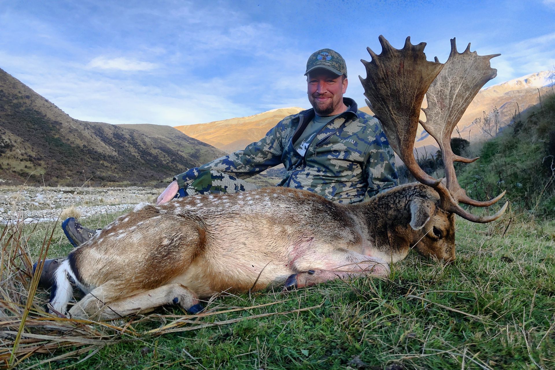 Hunter in camouflage sits beside a large, spotted deer with large antlers in a mountainous, grassy area.