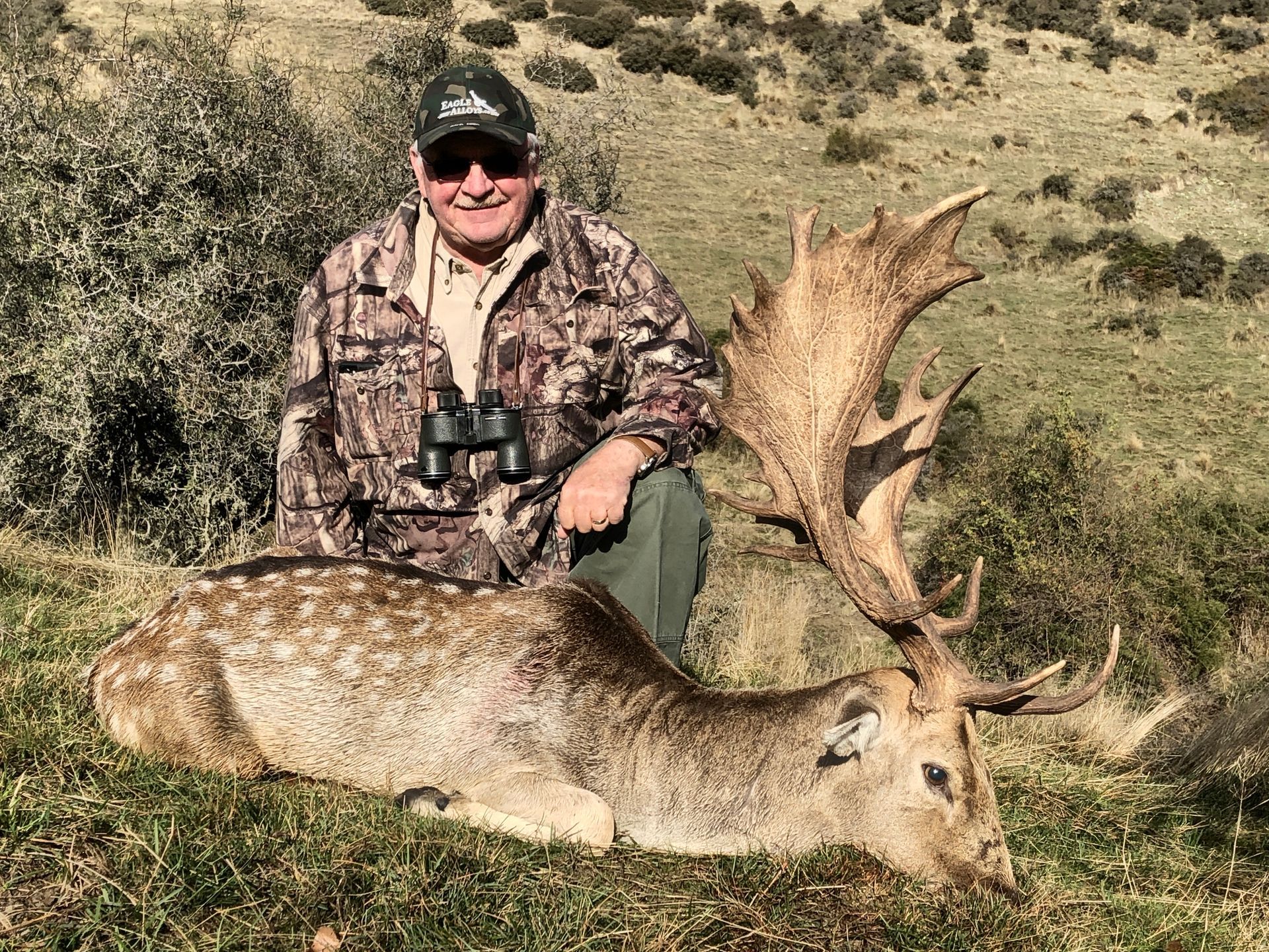 Man in camouflage with a large-antlered fallow deer he has apparently hunted. The setting is a grassy hill.