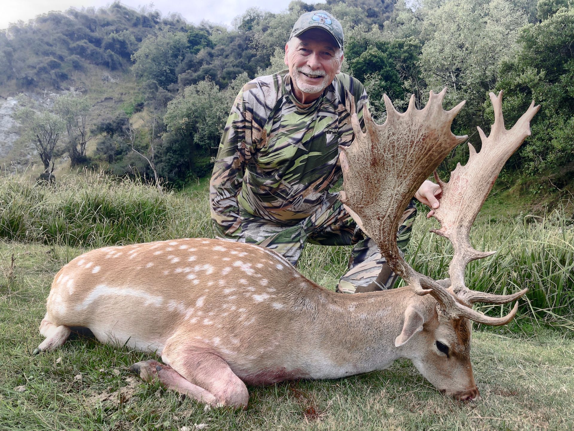 Man kneels beside a large dead fallow deer with impressive antlers. He smiles, dressed in camouflage, outdoors.