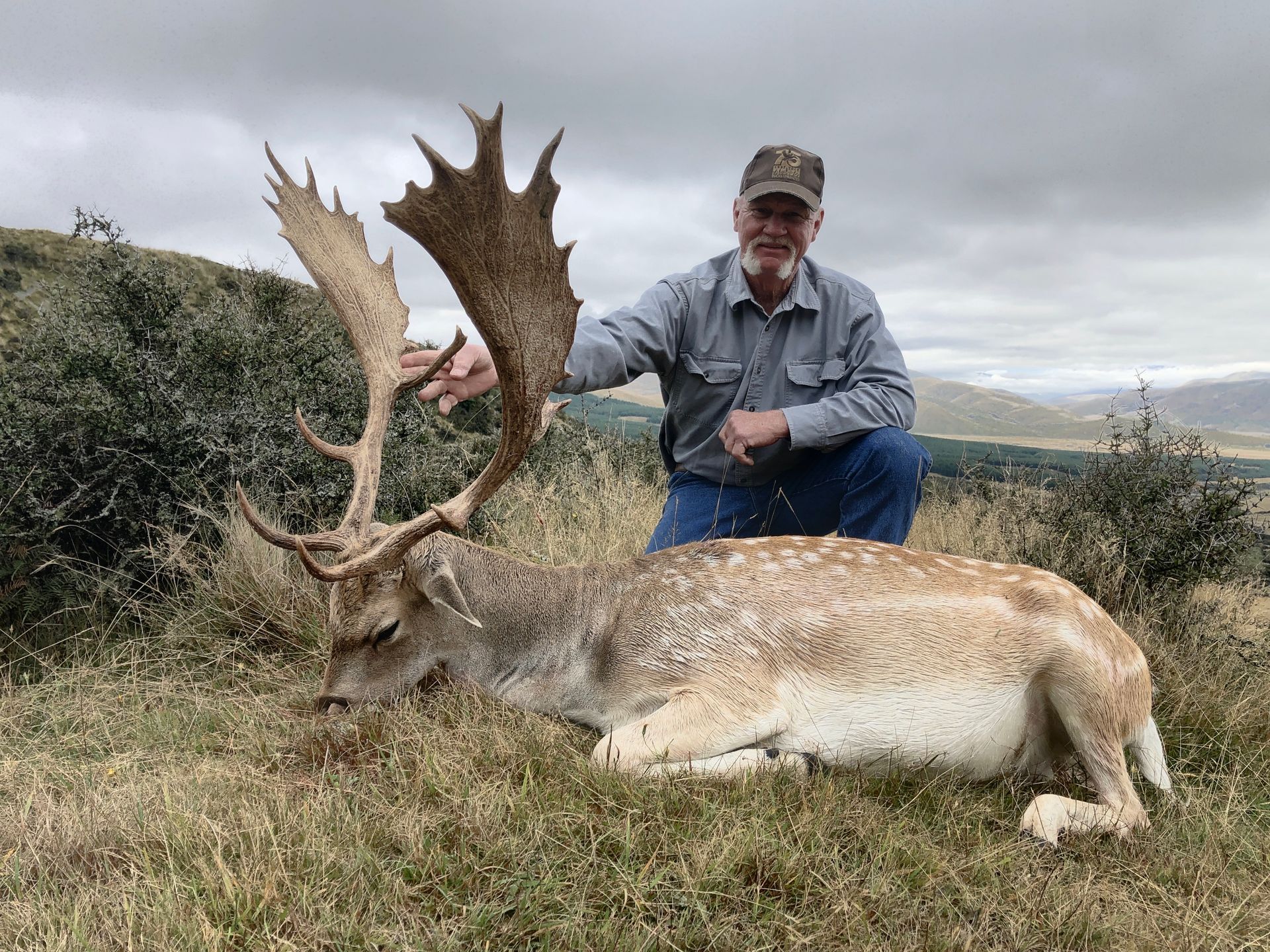 Man kneels next to a large, dead fallow deer with impressive antlers on a hillside. He is smiling.
