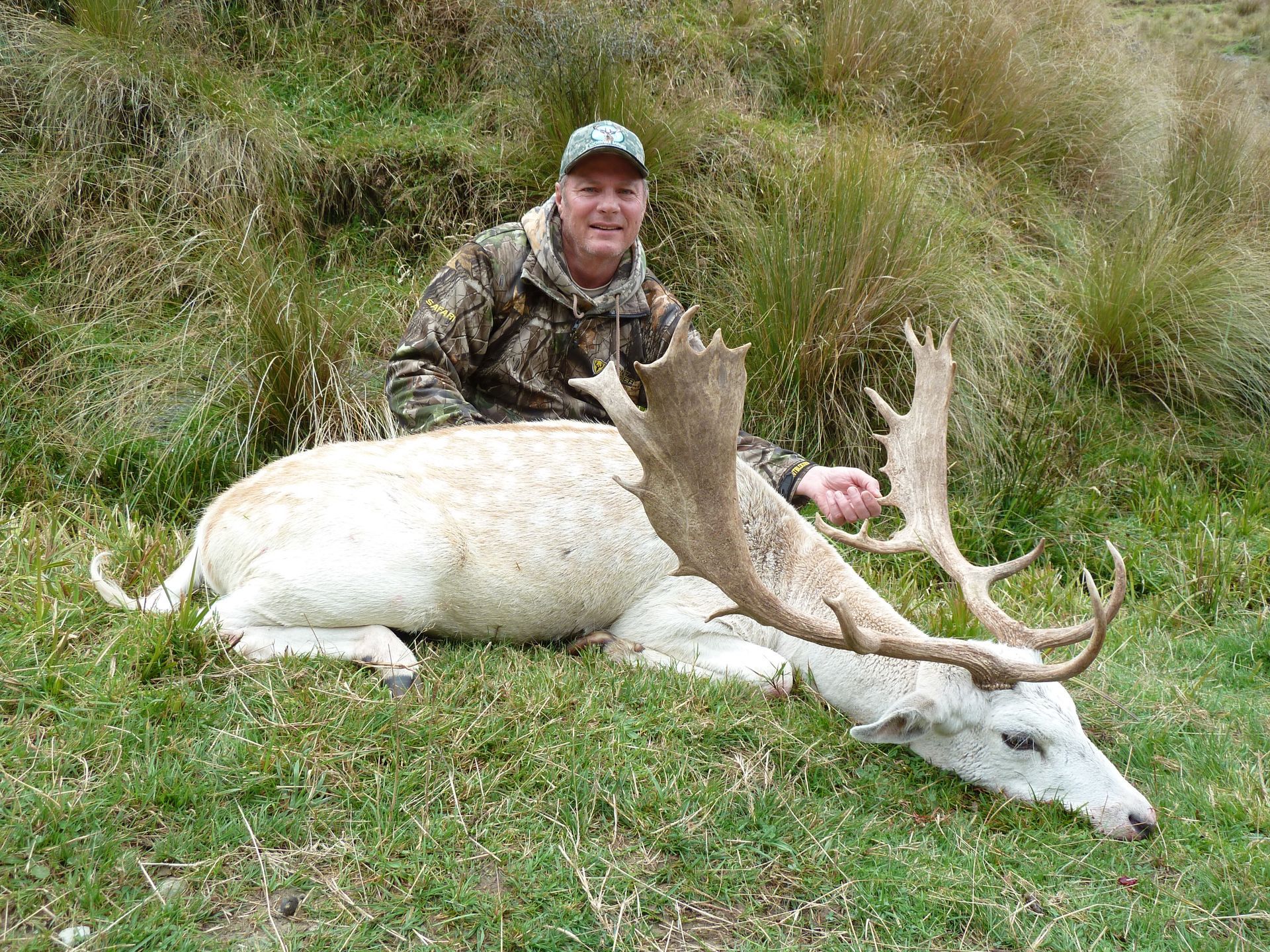 Man in camouflage with a large, white deer he has killed, resting on grass. He is smiling and gesturing toward the animal's antlers.