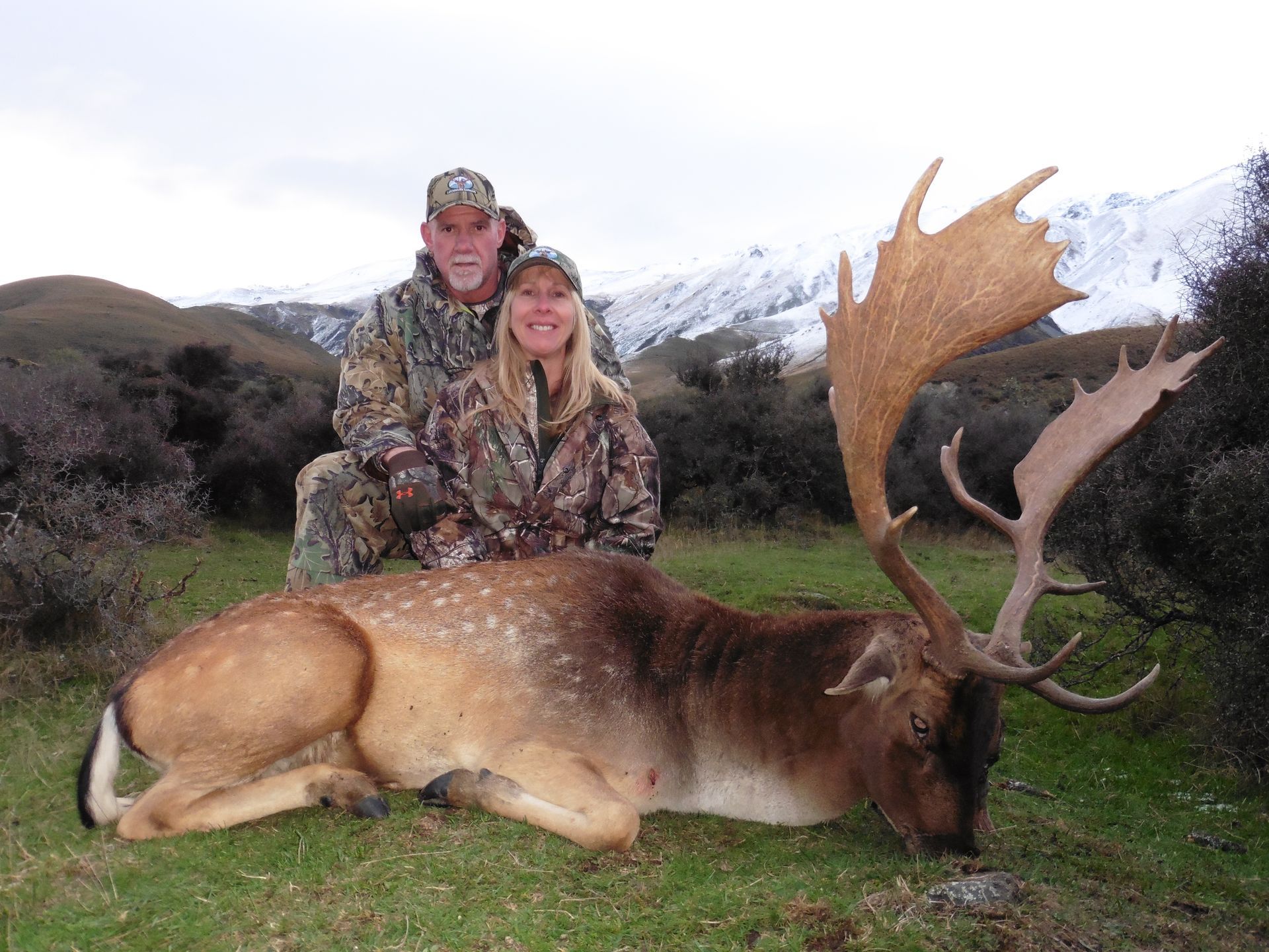 A man and woman in camouflage pose with a large deer they have hunted, in a grassy field with mountains in the background.