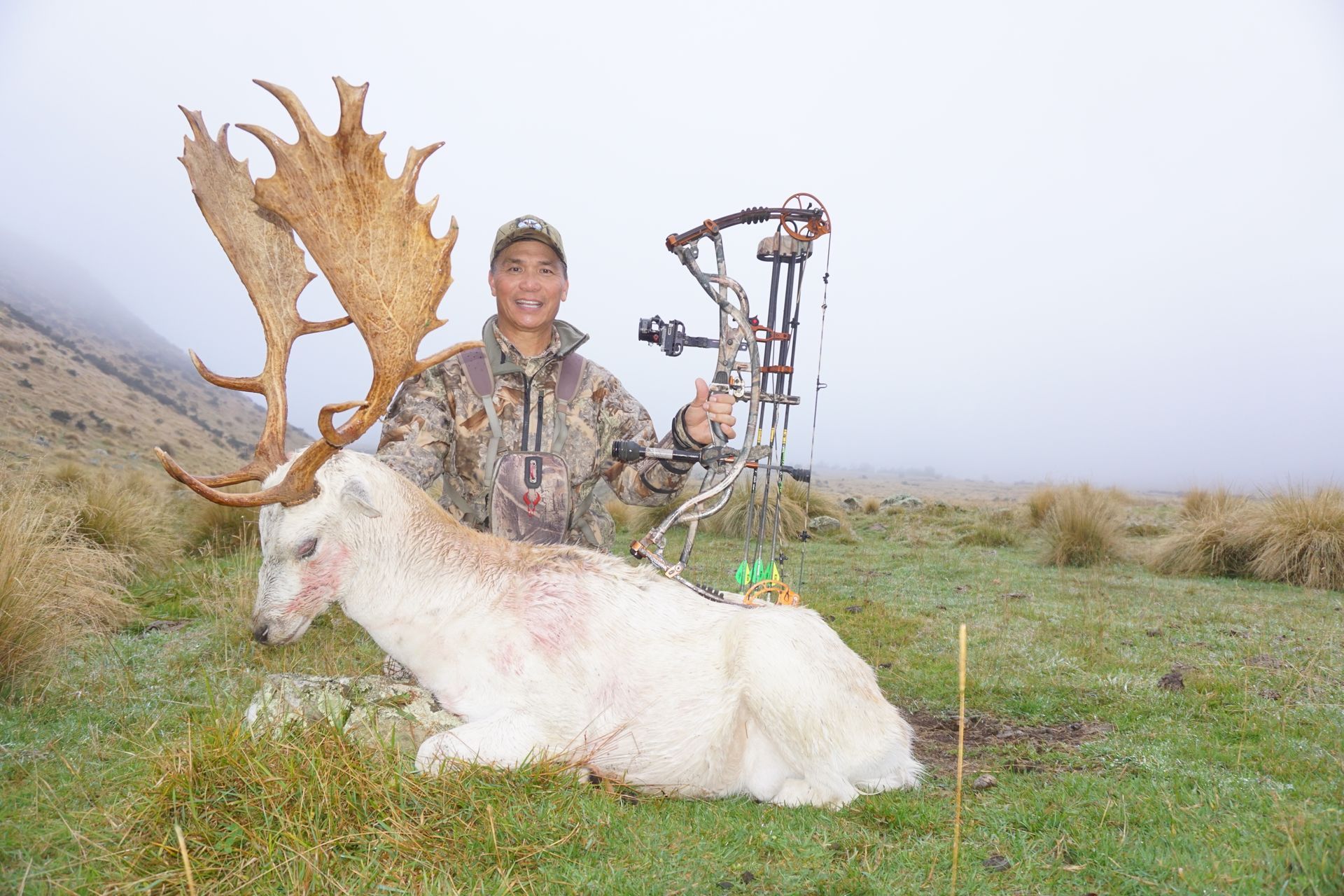 Hunter in camouflage smiles, posing with a large albino deer and bow in a grassy, foggy landscape.