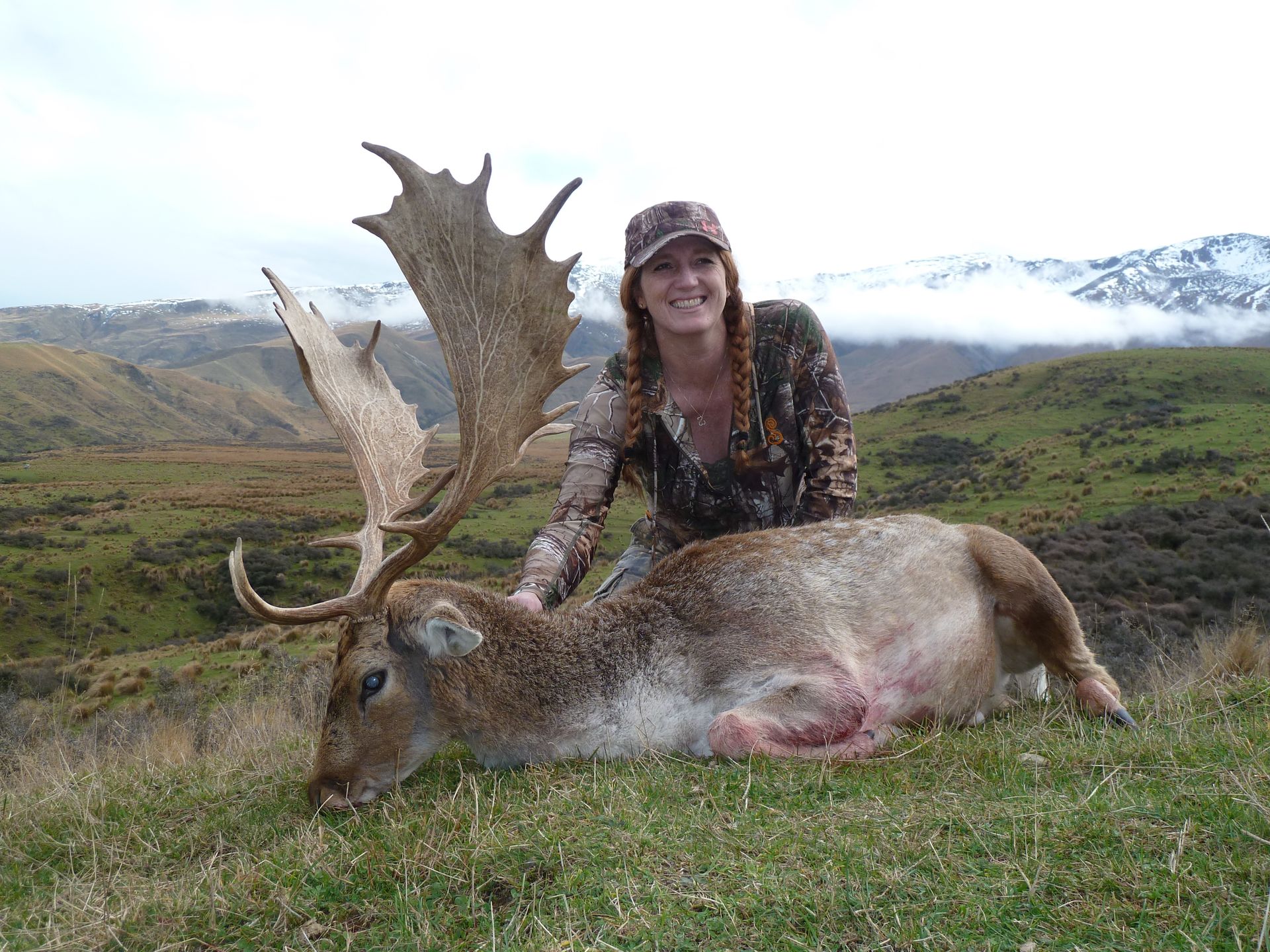 Woman in camouflage stands next to a dead deer with large antlers on a grassy hillside.