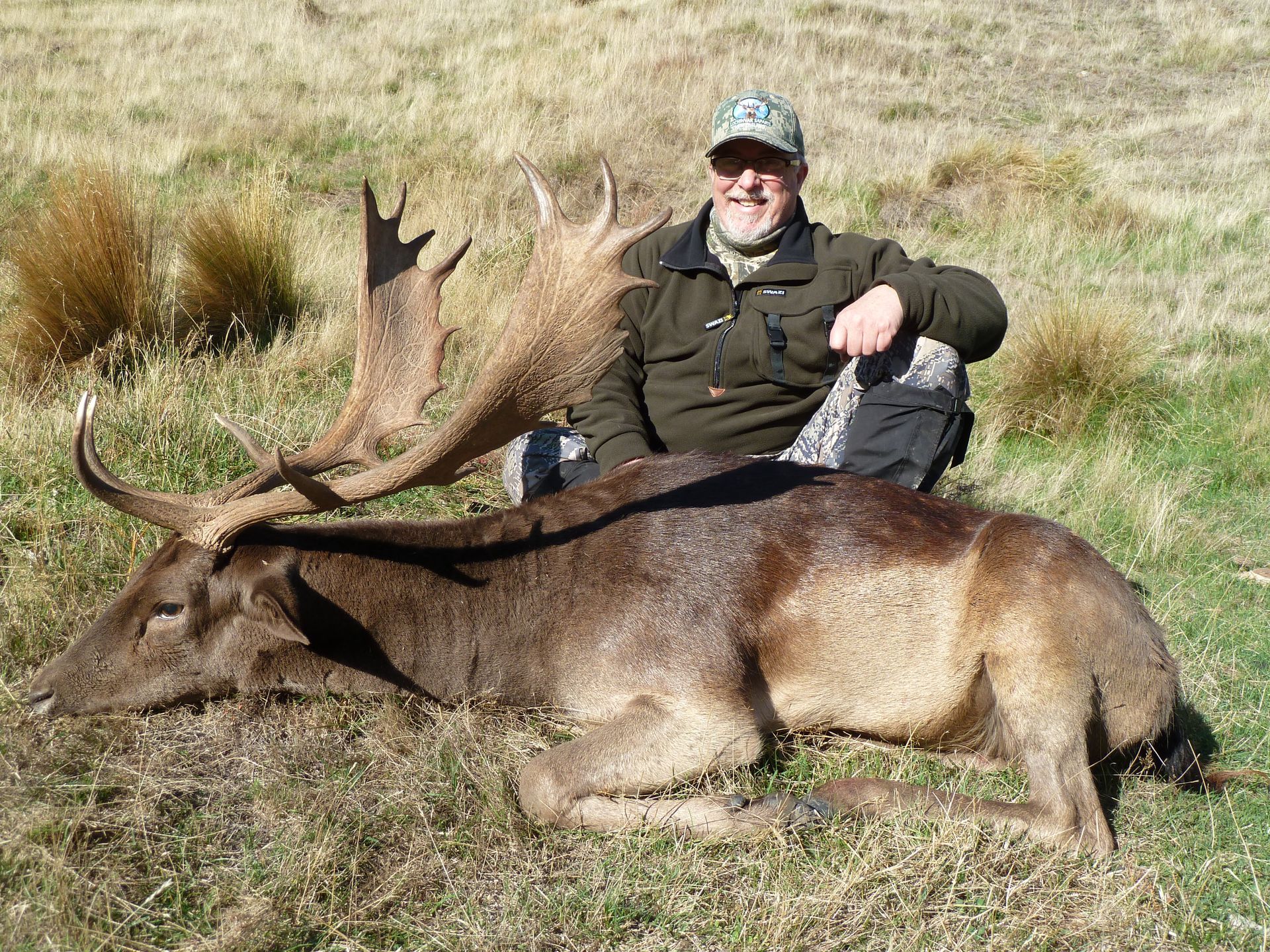 Man in hunting attire kneels beside a large deer with antlers on a grassy field. He's smiling.