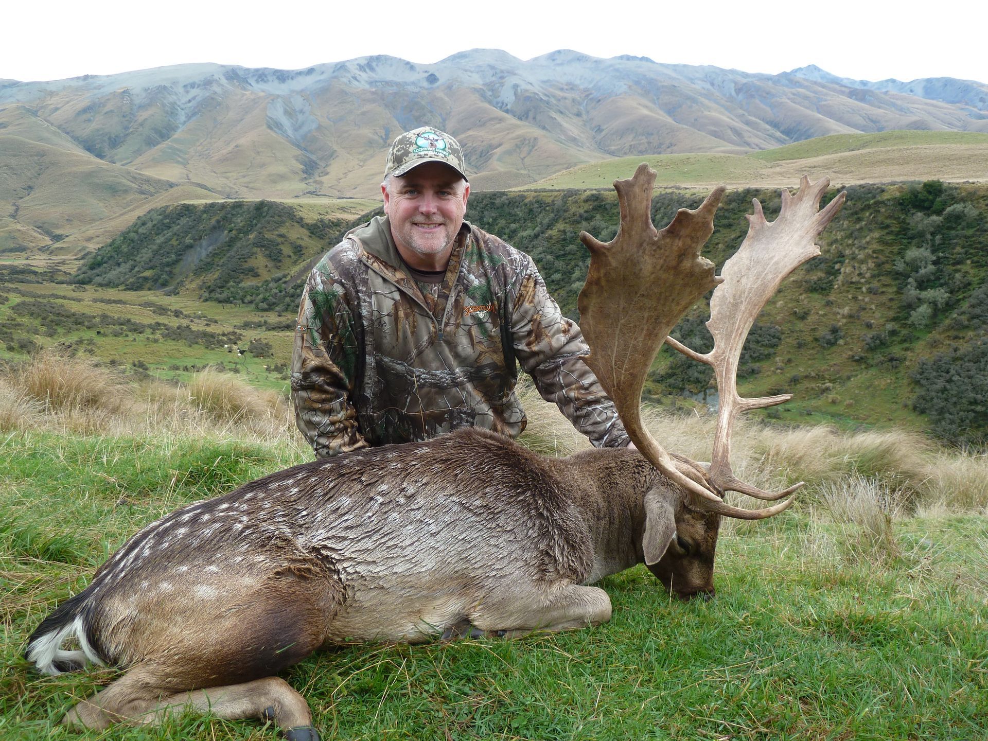 Man in camouflage gear with a large fallow deer he has hunted, posed in a grassy mountainous landscape.