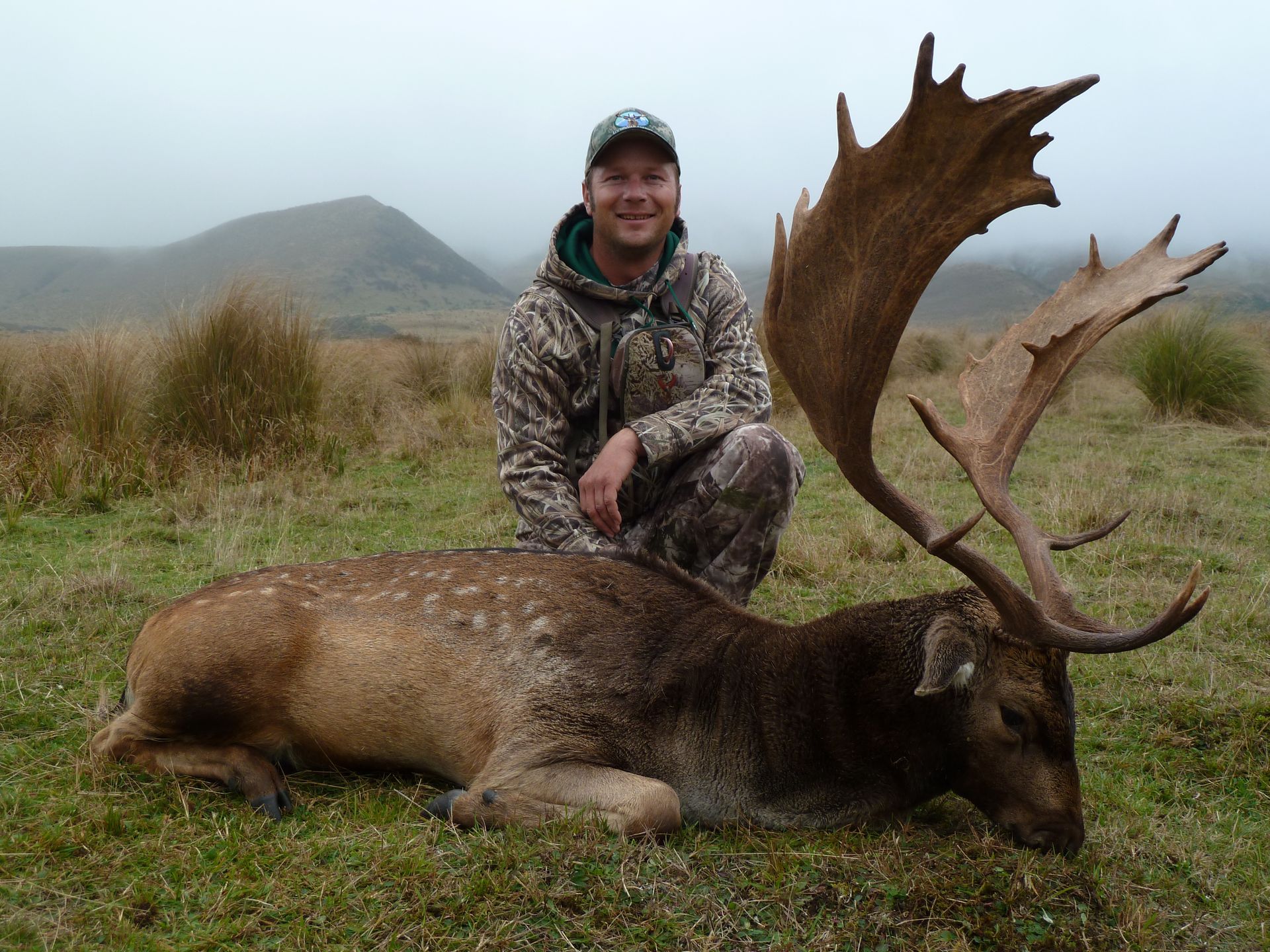 A man in camouflage kneels beside a large deer with impressive antlers in a grassy field on a foggy day.