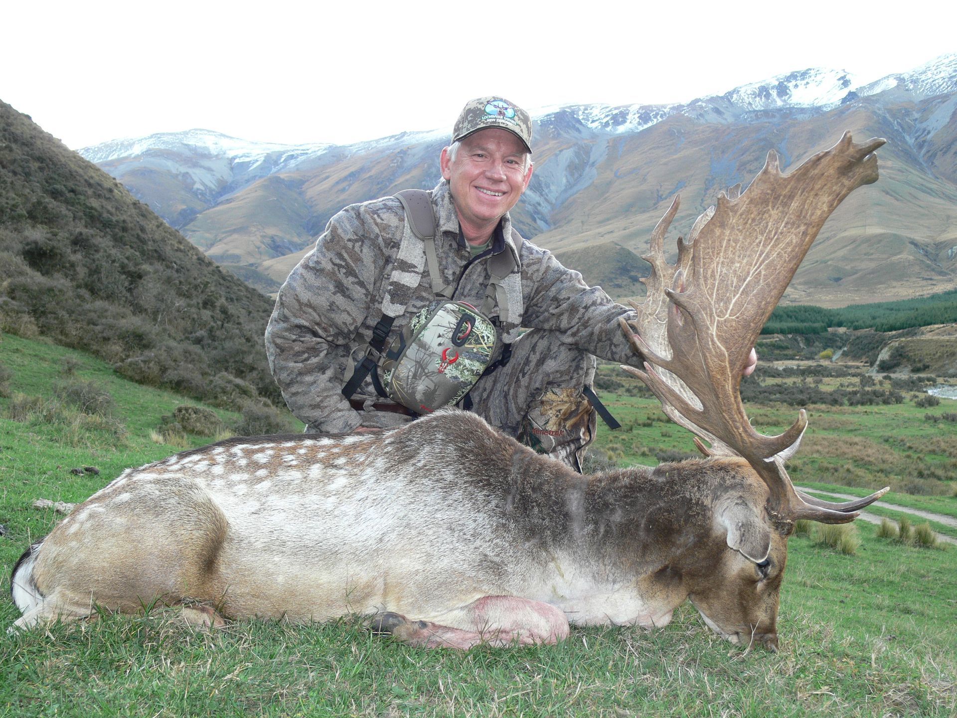 Man in camouflage kneeling beside a large dead deer with impressive antlers, against a backdrop of mountains.