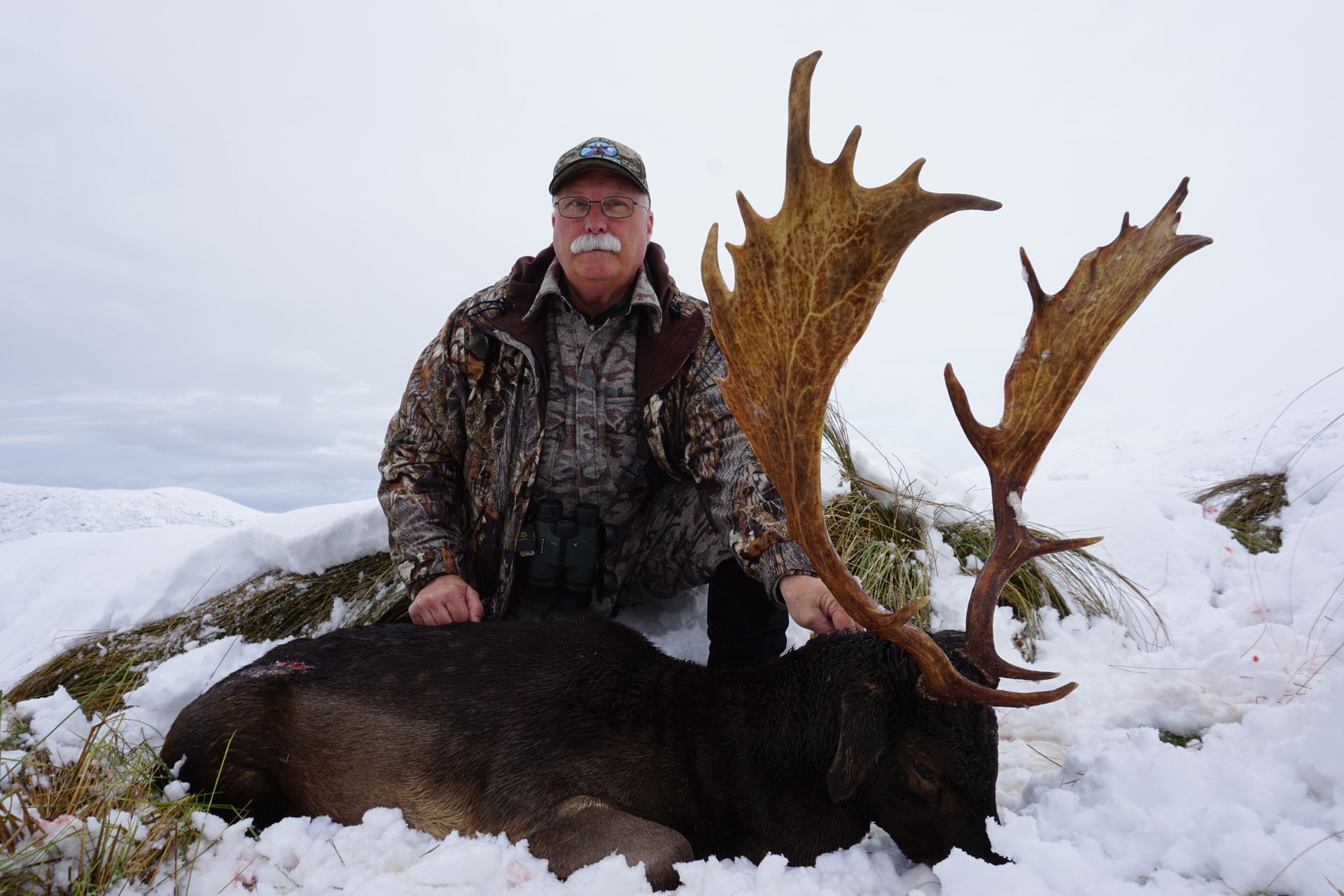 Man in camo jacket kneels beside a large, dead deer with impressive antlers in a snowy, outdoor setting.