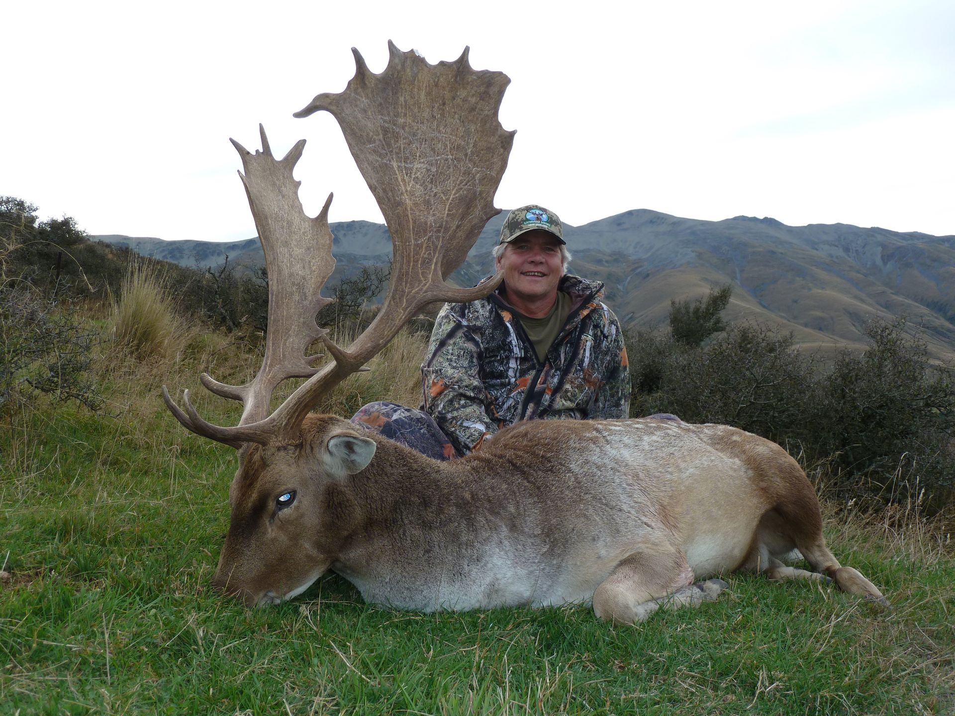 Man in camo gear smiles next to a large dead deer with impressive antlers in a grassy field with mountains in the background.