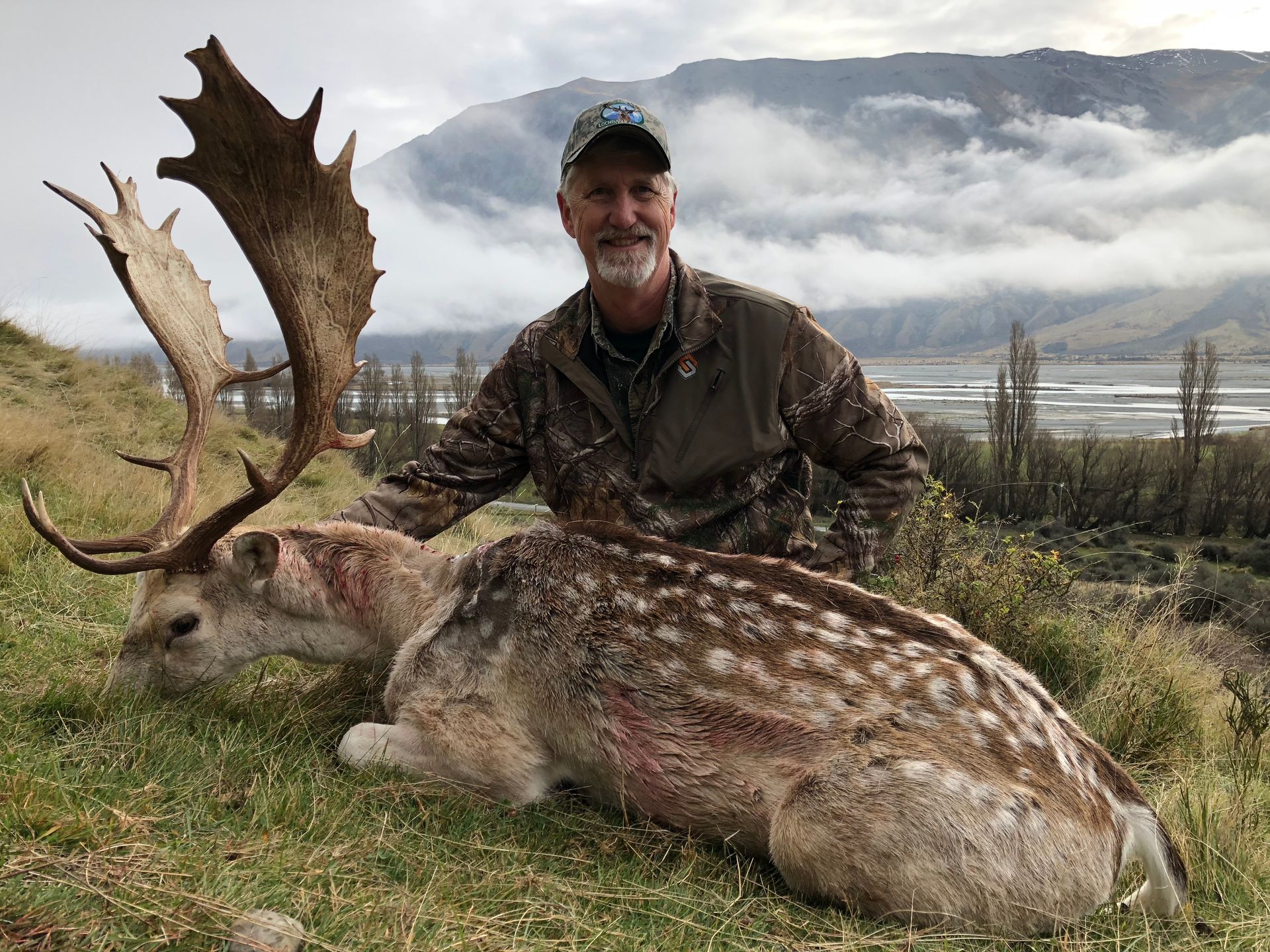 Man in camouflage with a large fallow deer he has killed. Deer has large antlers, and man is smiling in a mountainous landscape.