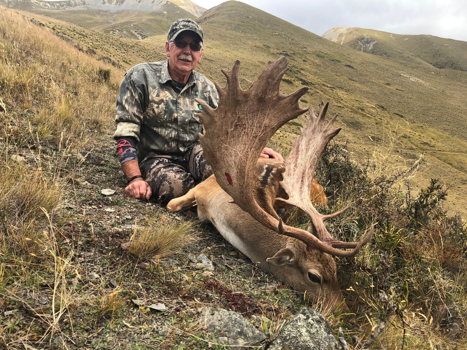 A hunter kneels beside a dead deer with large antlers on a hillside. He wears camouflage and smiles.