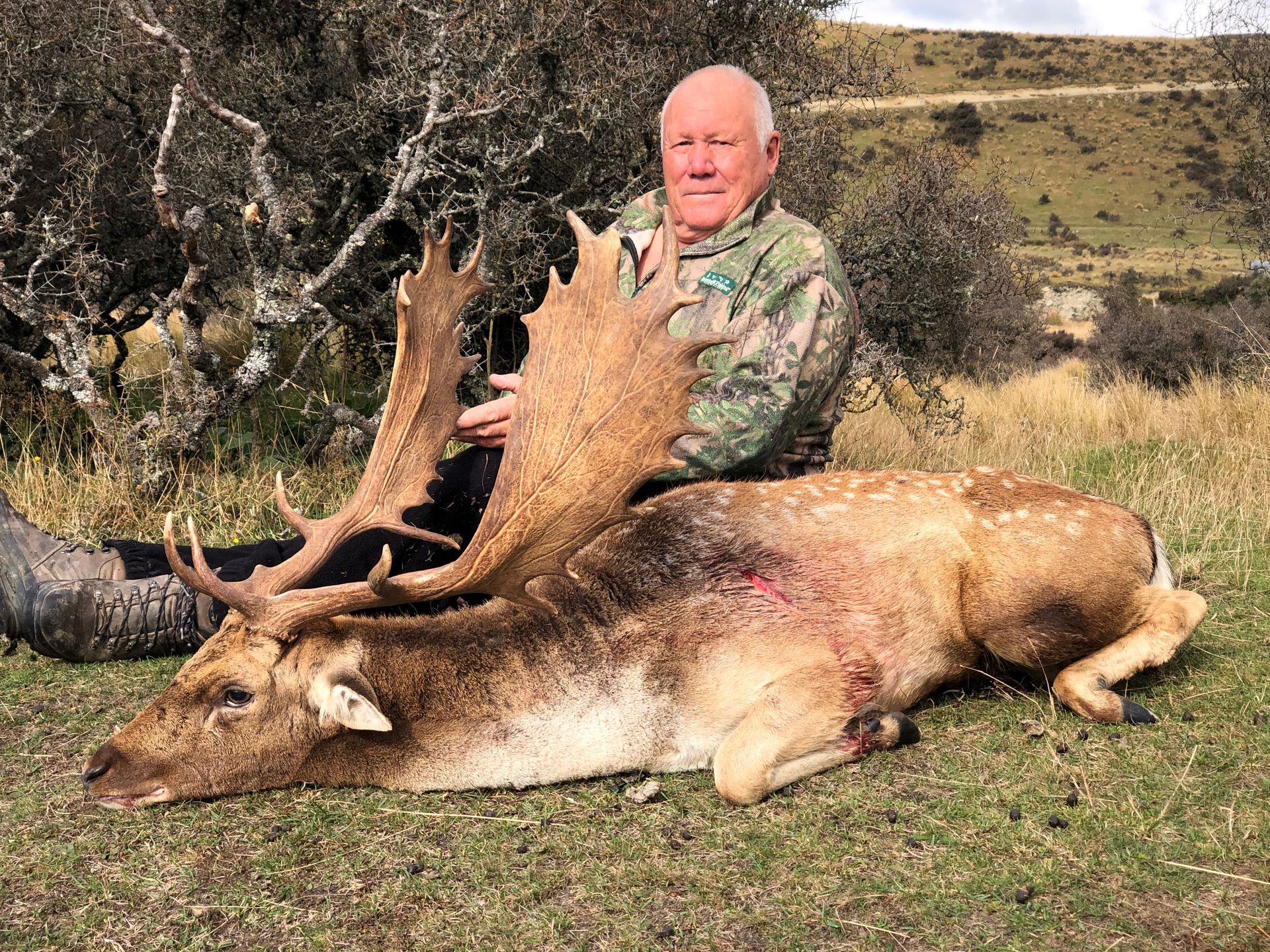 Man in camouflage posing with a large, dead deer with impressive antlers in a grassy field.