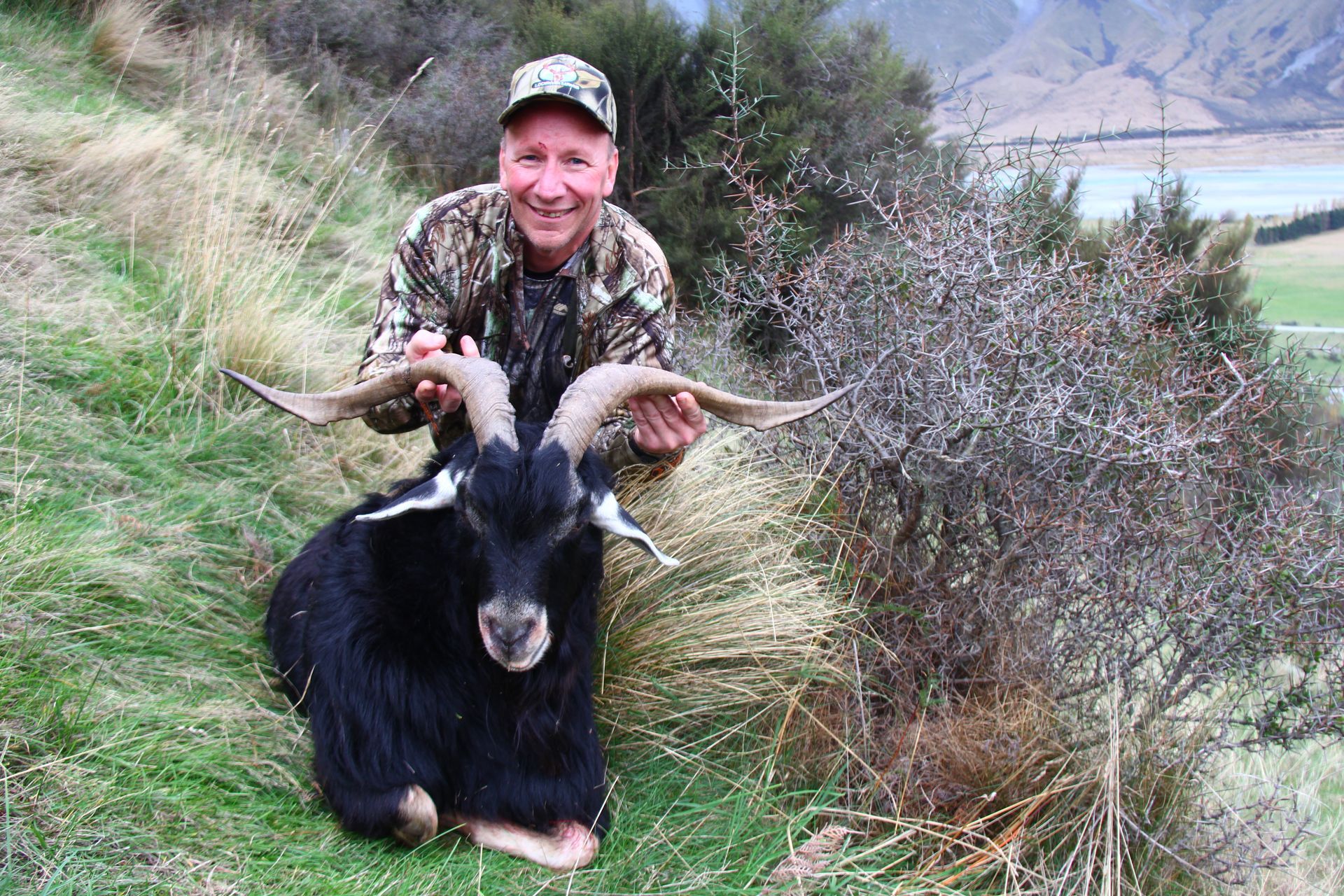 Man in camouflage holding a black goat with large horns, smiling in a grassy hillside setting.