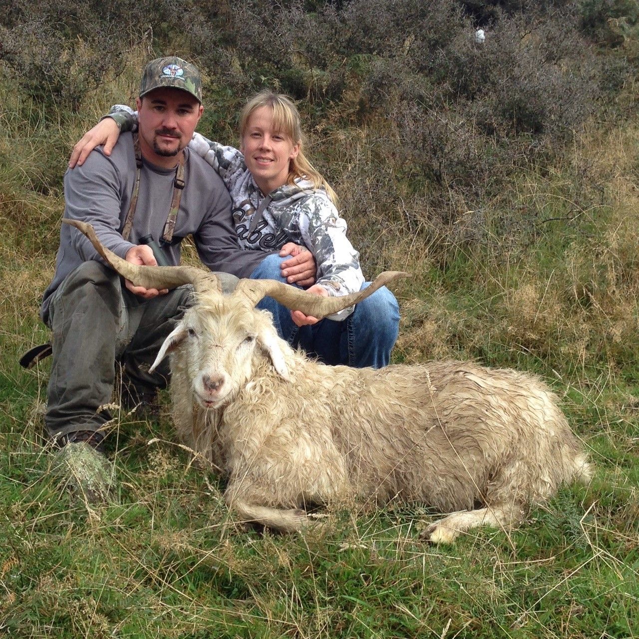 A man and a girl pose with a goat they hunted. The goat is lying on the ground in tall grass. They are in an outdoor setting.