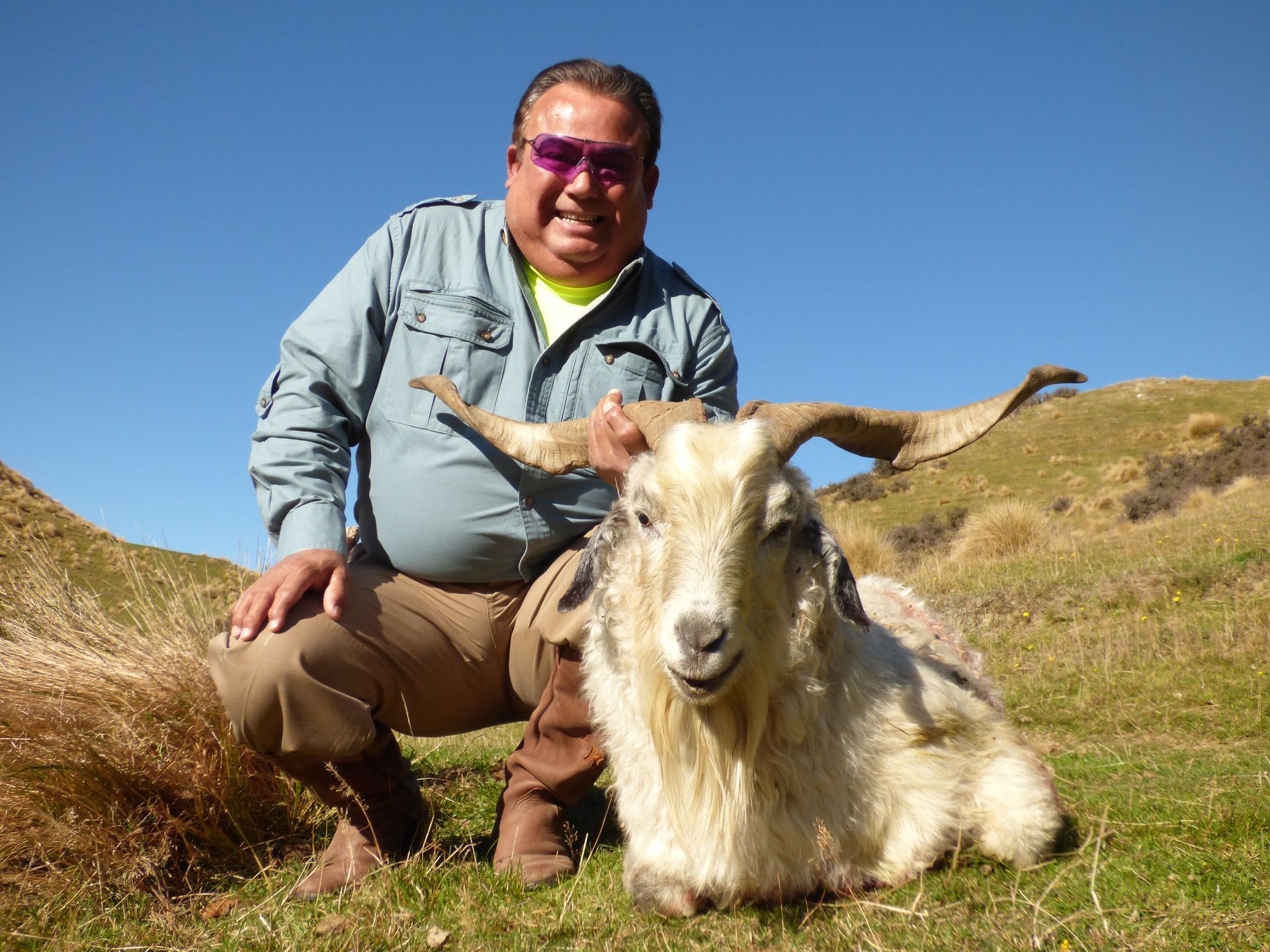 Man in sunglasses kneels beside a large goat with long horns. They are in a grassy field with a blue sky.