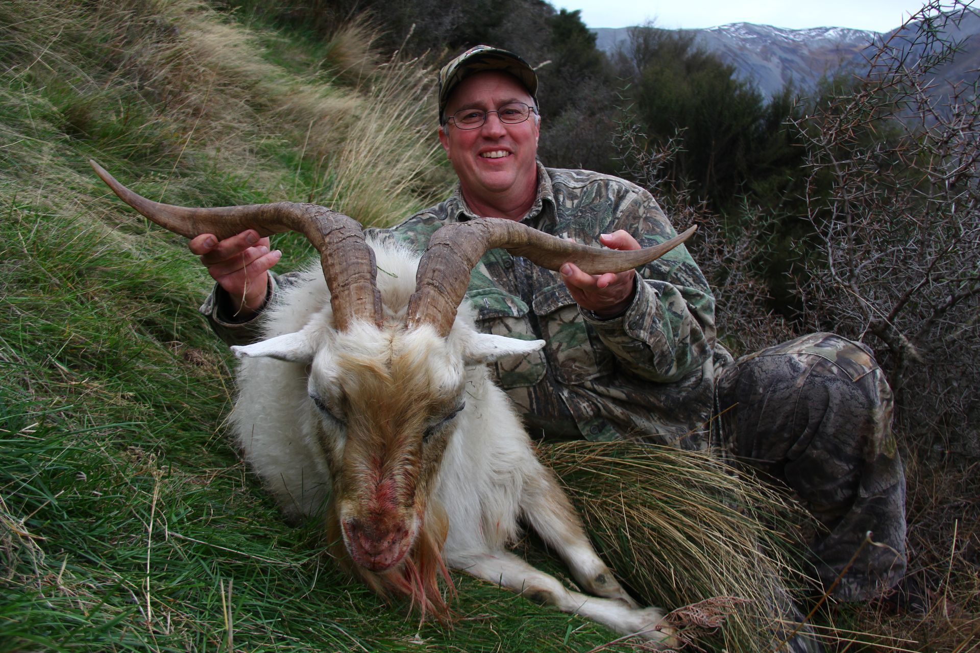 Man in camouflage holds up the head of a dead, large goat with curved horns. He is kneeling in grass.