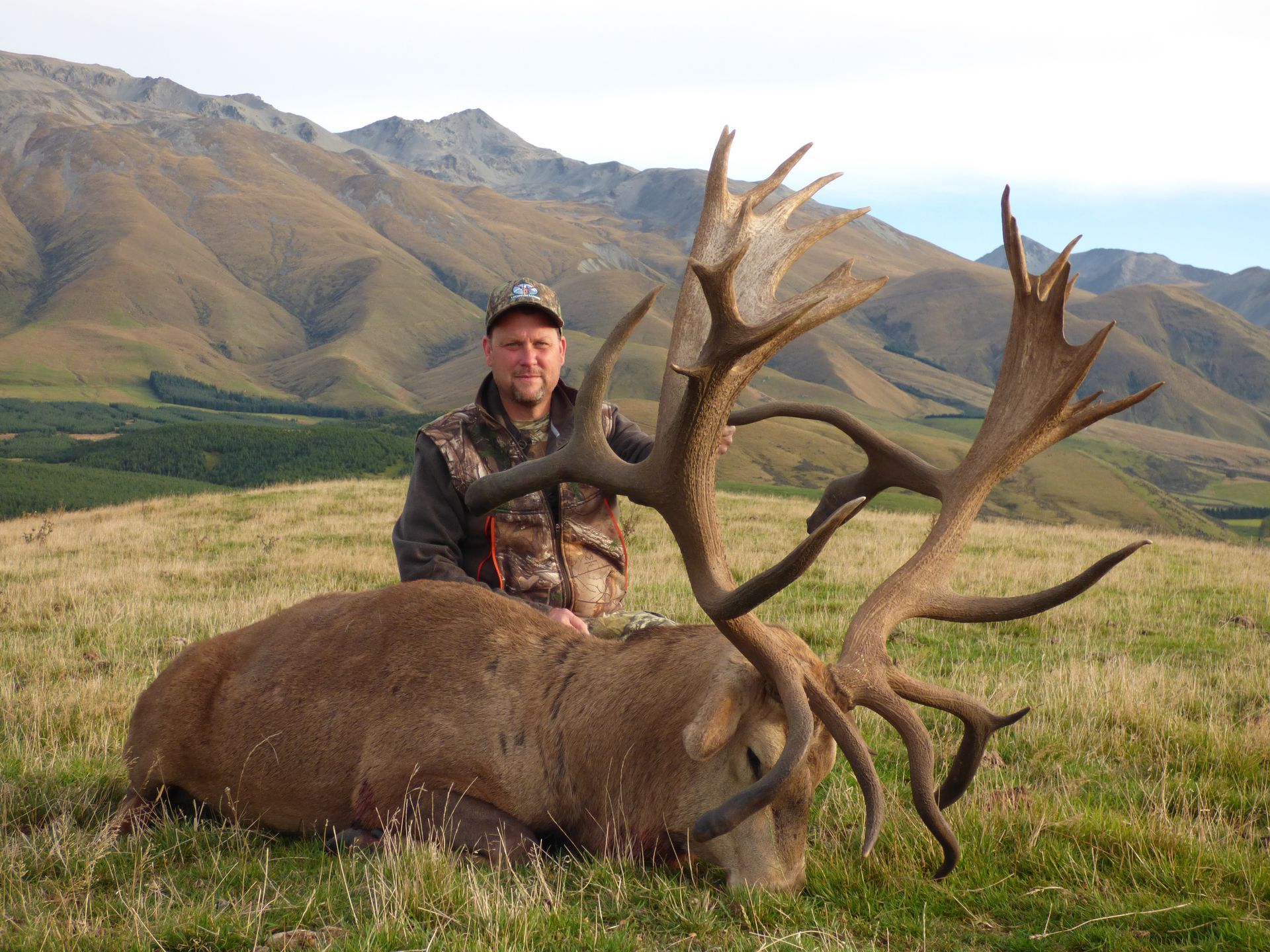 Hunter with a large dead deer, posing in a grassy field with mountains in the background.
