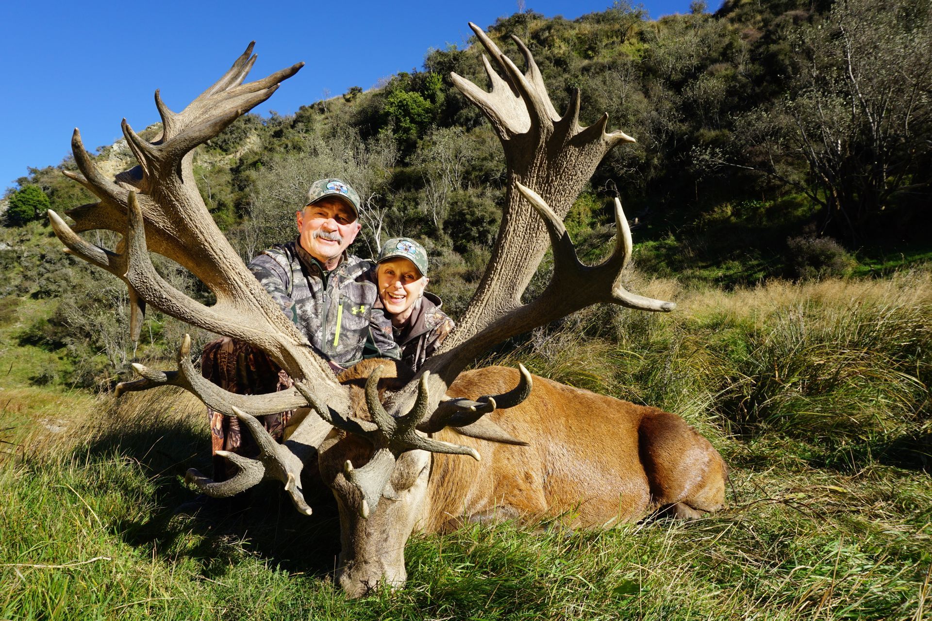 Two people pose with a large dead red deer, holding its antlers. They're smiling outside in green grass, hills in the background.