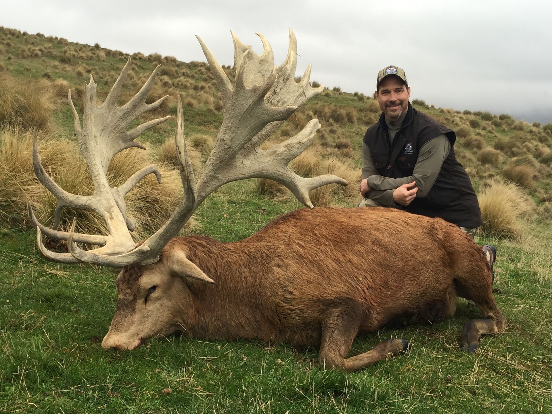 A man smiles beside a large dead red deer with enormous antlers on a grassy hillside.