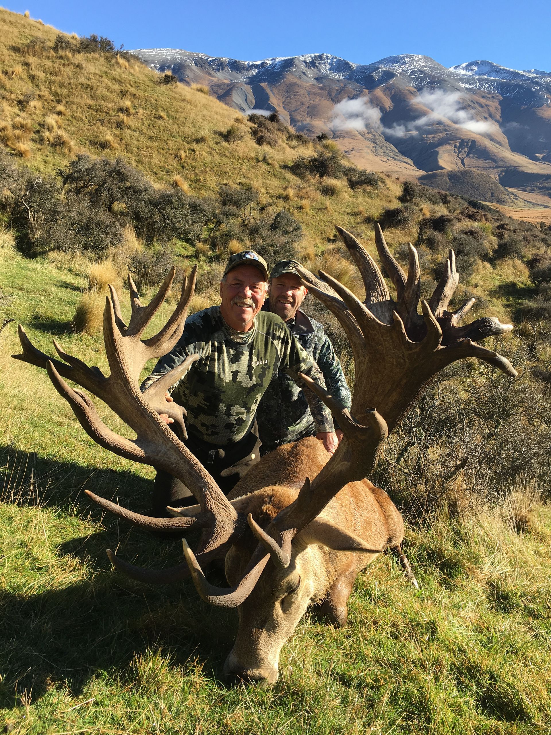 Two men smiling, posing with a large deer they've hunted in a mountainous, grassy area. The deer has large antlers.