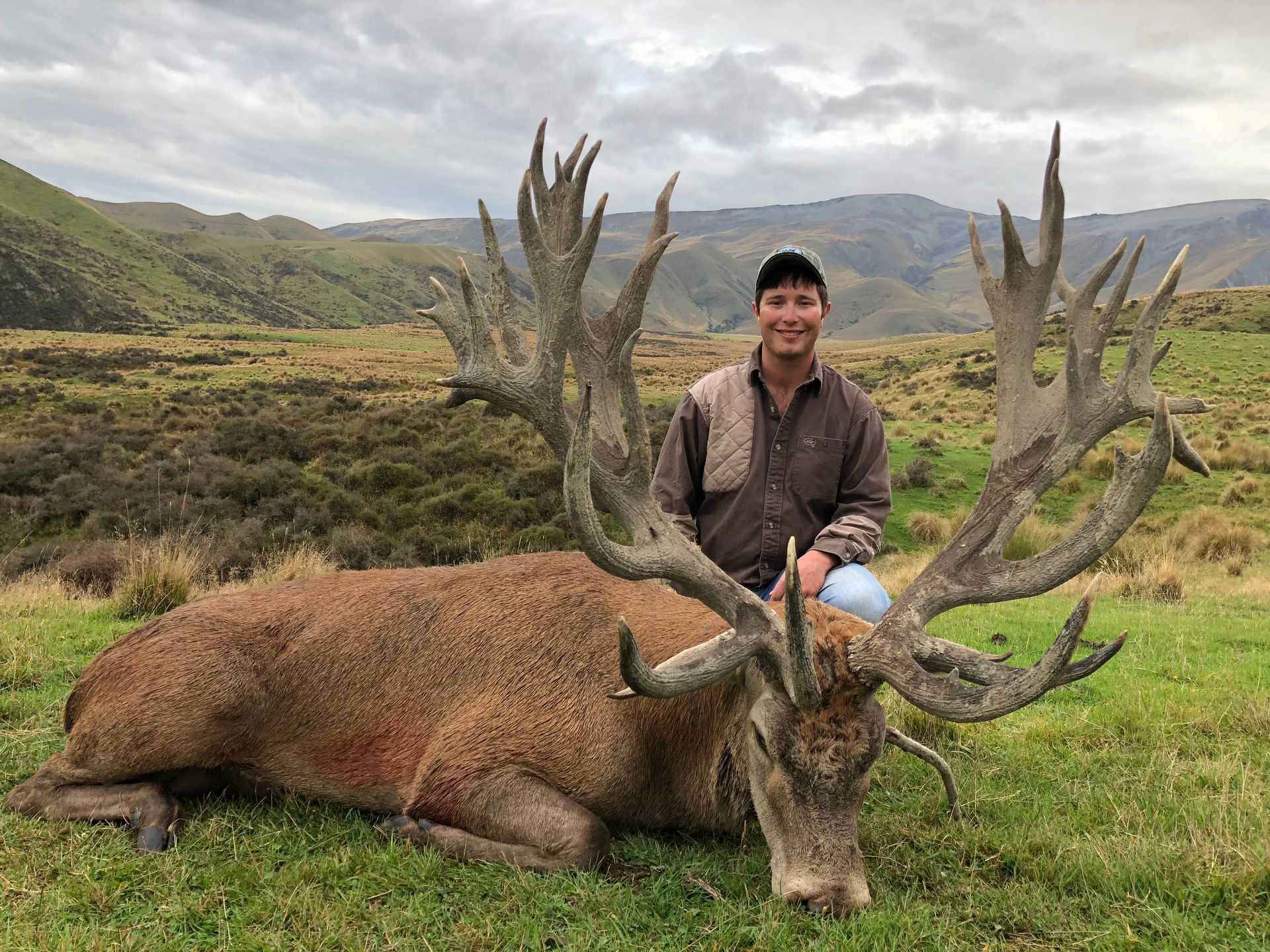 Man smiles next to a large red deer lying on grass. The deer has massive antlers and a brown coat; mountains are in the background.