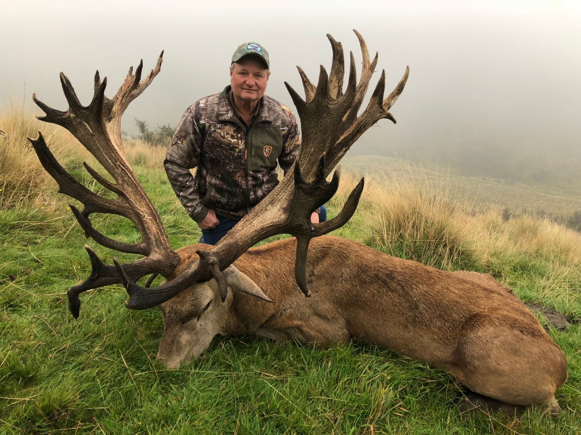 Man in camouflage poses with a large dead red deer on a grassy hillside. The deer has massive antlers.